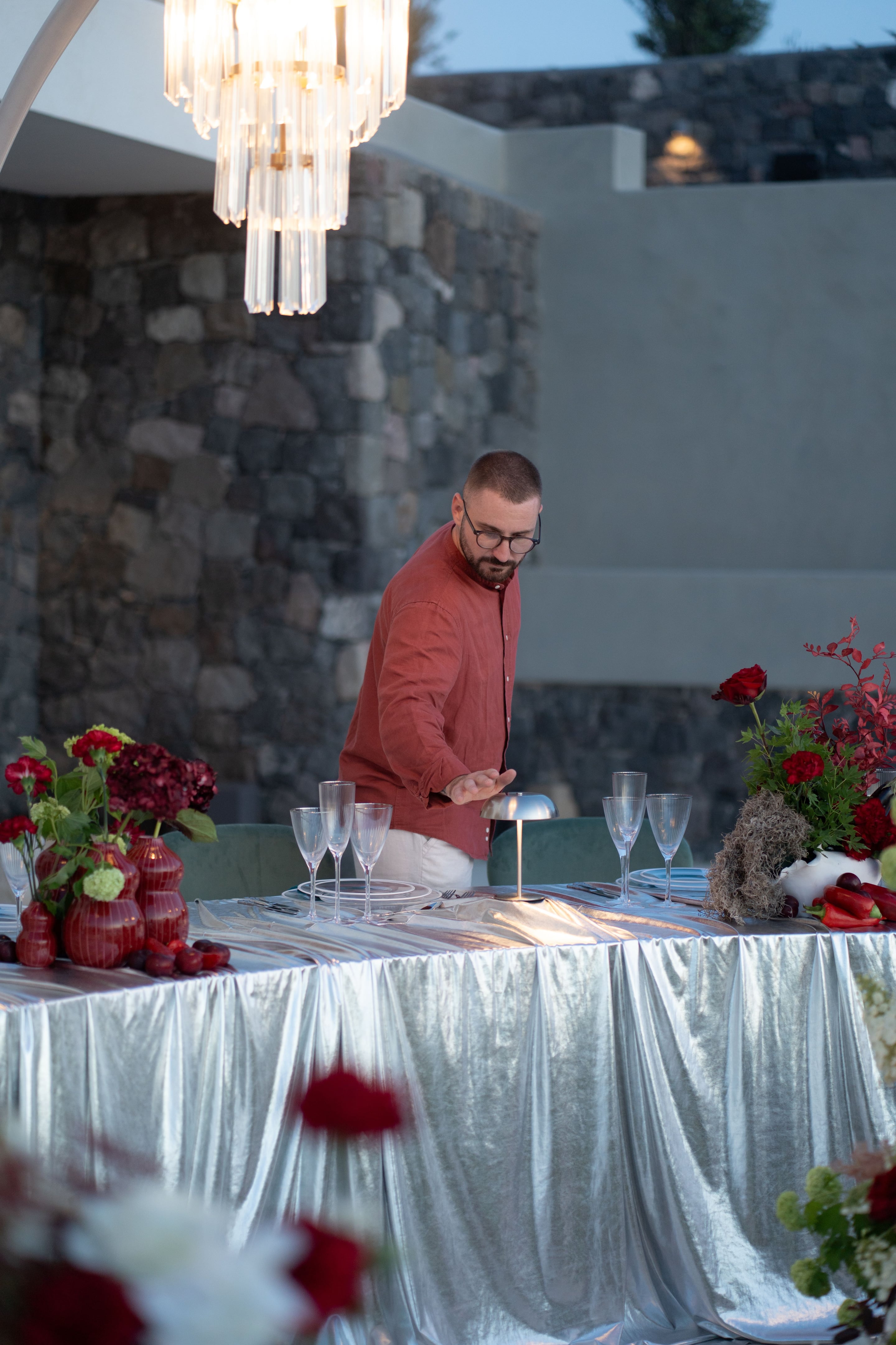 A bearded man in glasses and a rust-red linen shirt leans over a long table draped in shimmering silver fabric, adjusting a small silver table lamp among crystal wine glasses and white plates. At the table's edge, deep red garden roses, dark burgundy hydrangeas, and sprays of red foliage fill red ceramic vases and a white bowl, with bright red peppers and plums scattered nearby. This is a wedding table setting at Islet in Santorini, Greece, with a rough stone wall and soft evening light behind.