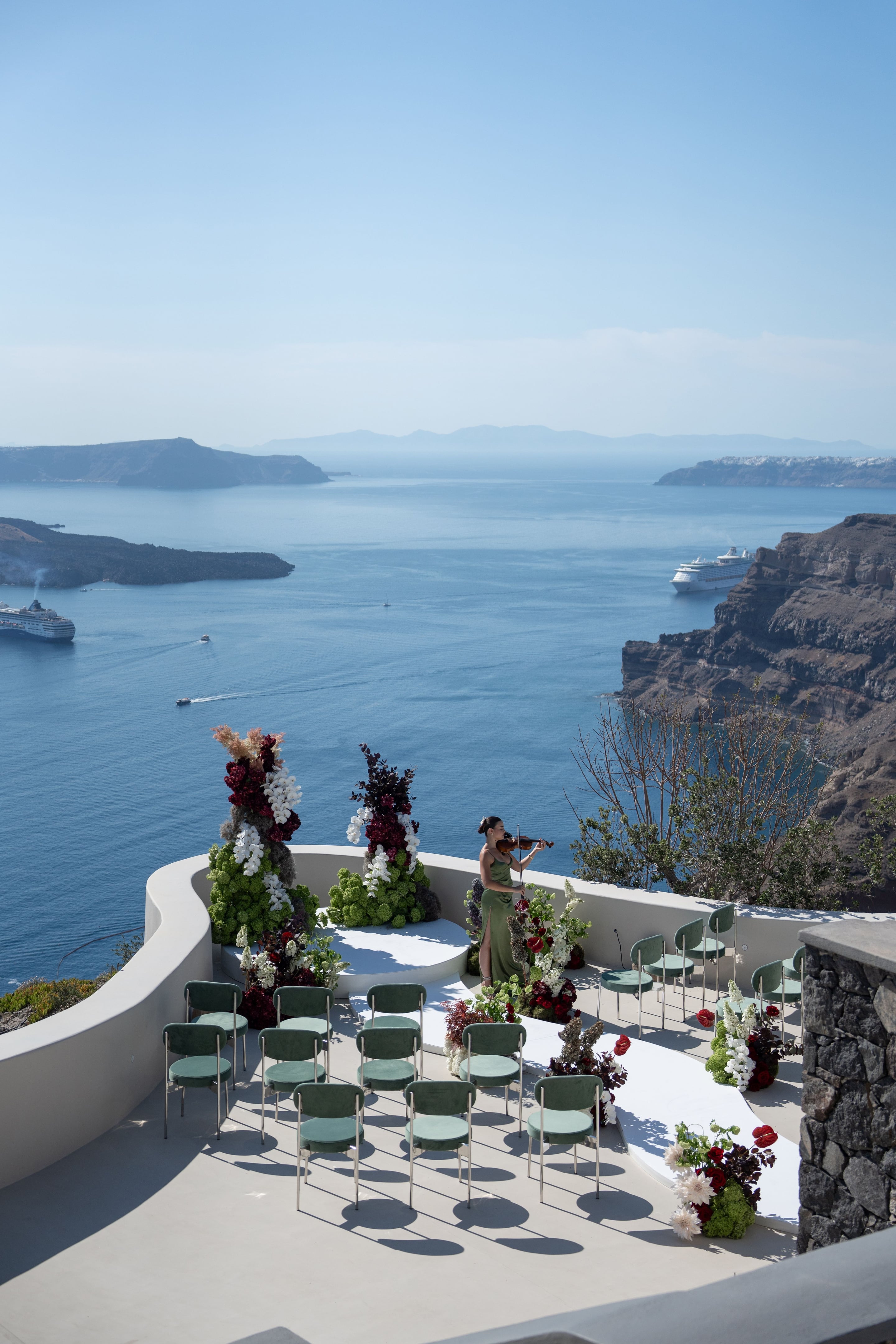 A wedding ceremony set on a whitewashed terrace above the caldera at Islet in Santorini, Greece, with the blue sea far below. Two tall floral towers of green hydrangea-like blooms, white orchids, deep burgundy foliage and dried pampas stand on a curved white platform, with a violinist in a green gown playing between them. Rows of sage-green velvet chairs face the aisle, which is lined with red anthurium, white dahlias and dark burgundy blooms, and cruise ships drift on the water beyond.