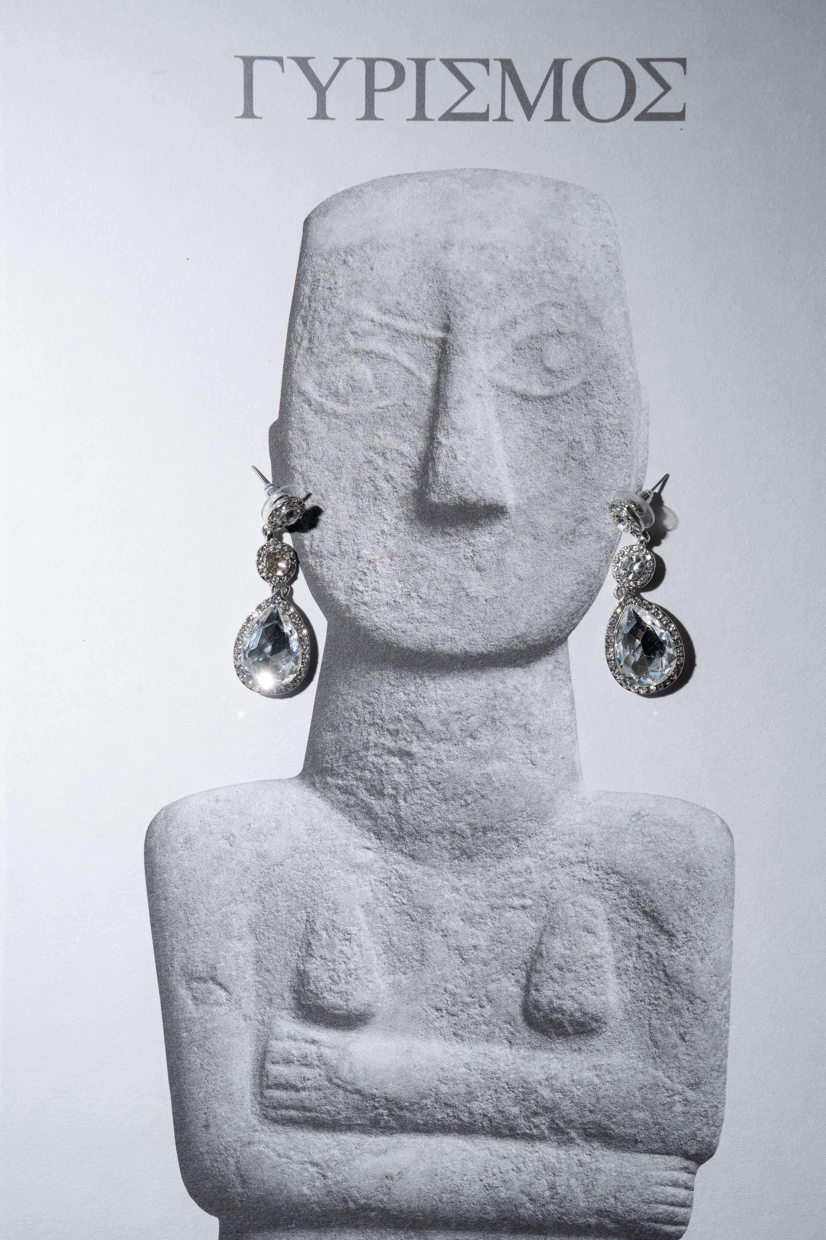 Close view of a grey, grainy photograph of a Cycladic-style marble figurine, its arms folded across the chest, with a pair of sparkling teardrop crystal earrings with pavé halos hanging from its ears against a pale background. The earrings are the only colour-bright element in the image. This is a wedding detail from Islet in Santorini, Greece, shared as part of Ellen and Jeff's story.
