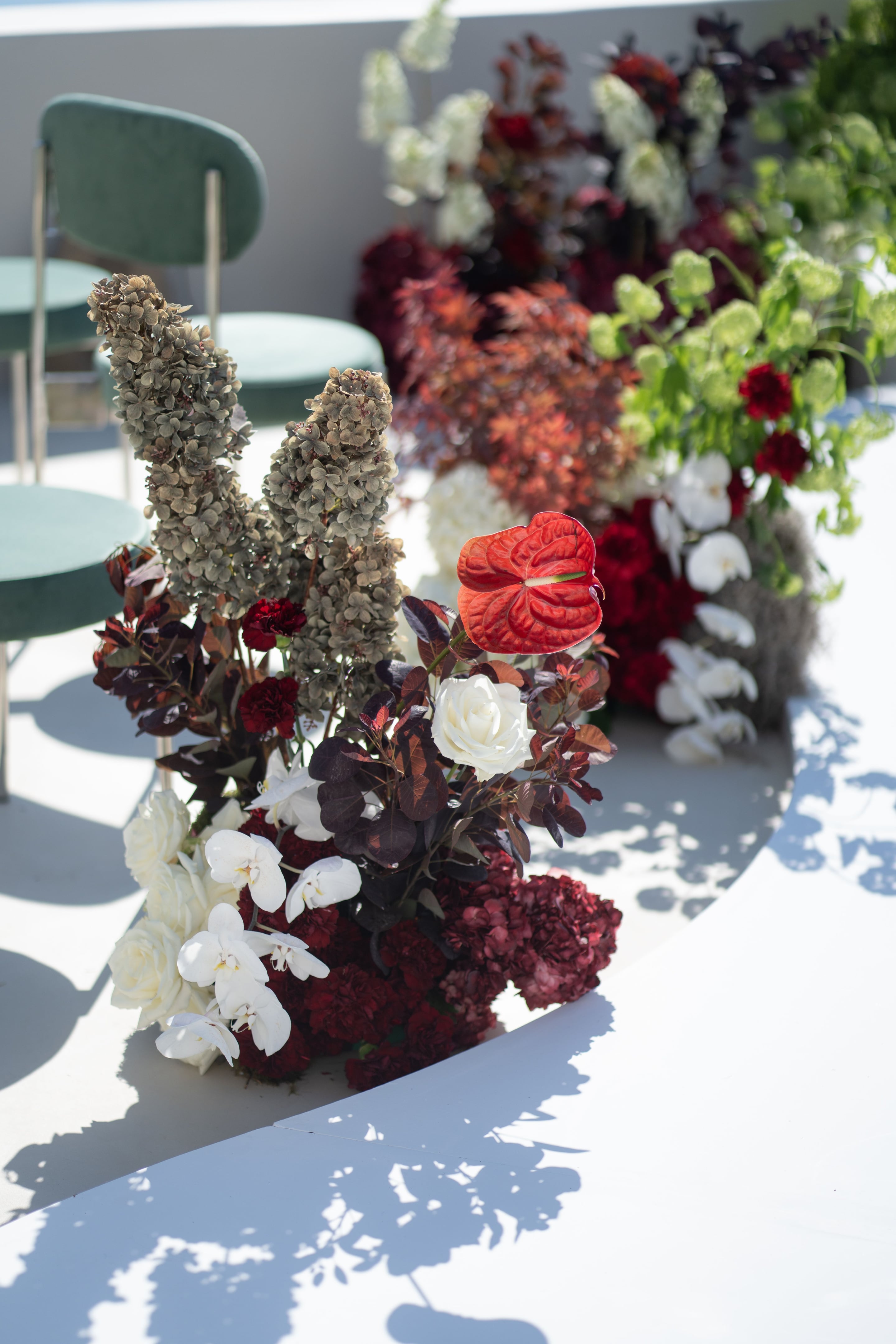 A wedding floral arrangement on a sunlit white terrace at Islet Santorini, Greece, featuring a bright red anthurium at the centre, surrounded by white roses, white phalaenopsis orchids, and deep burgundy carnations and hydrangeas. Dried silver-grey hydrangea stems rise on the left, with dark plum foliage woven through, while mint-green velvet chairs and blurred bursts of red and white blooms sit in the background.