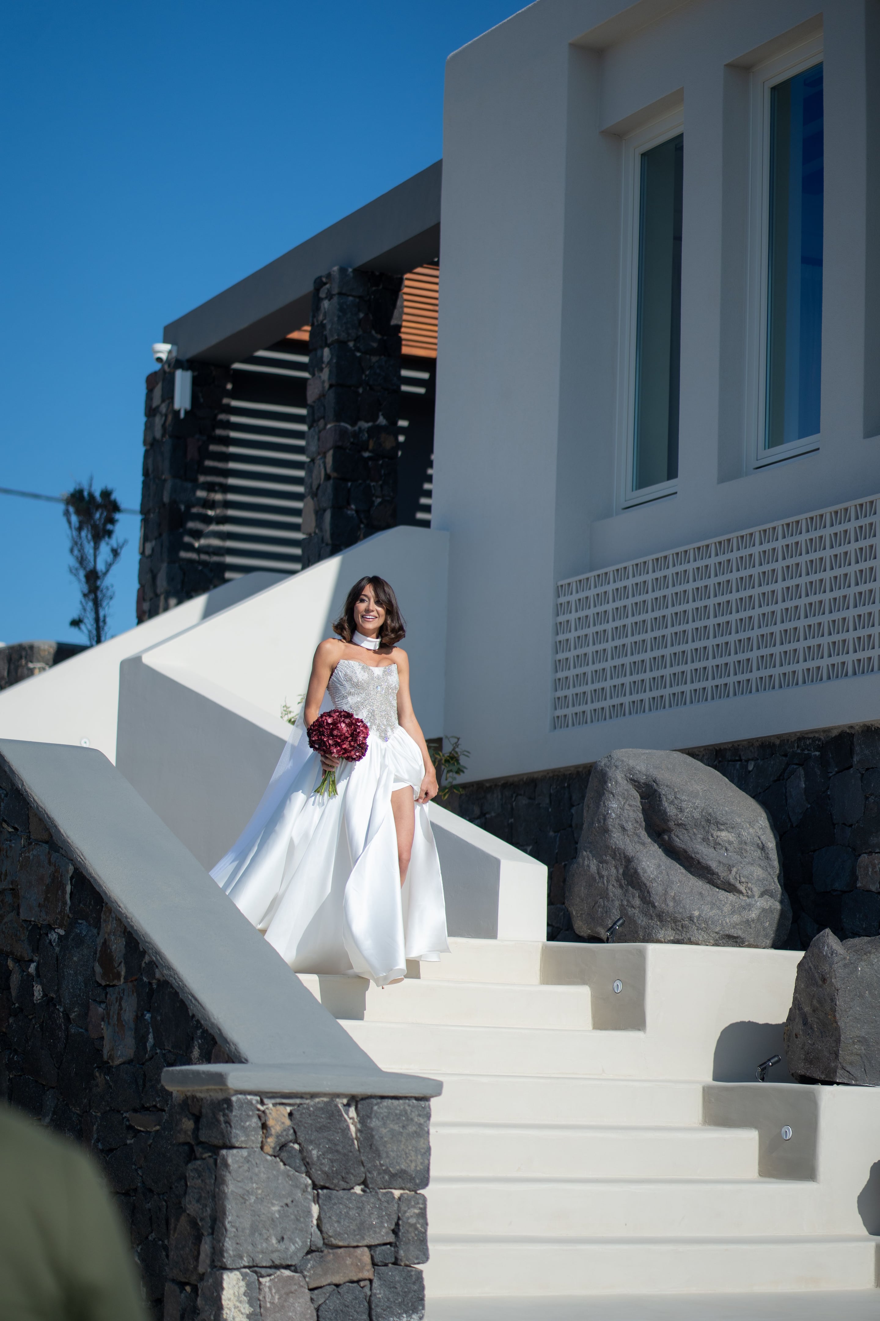 A bride in a white satin gown with a sparkling crystal-embellished bodice and a high white choker descends a whitewashed staircase, lifting her skirt to one side as she smiles. She holds a bouquet of deep burgundy hydrangea, with dark volcanic stone walls and large grey boulders framing the steps. This is a wedding bridal bouquet photographed at Islet Santorini in Greece, with bright blue sky above.