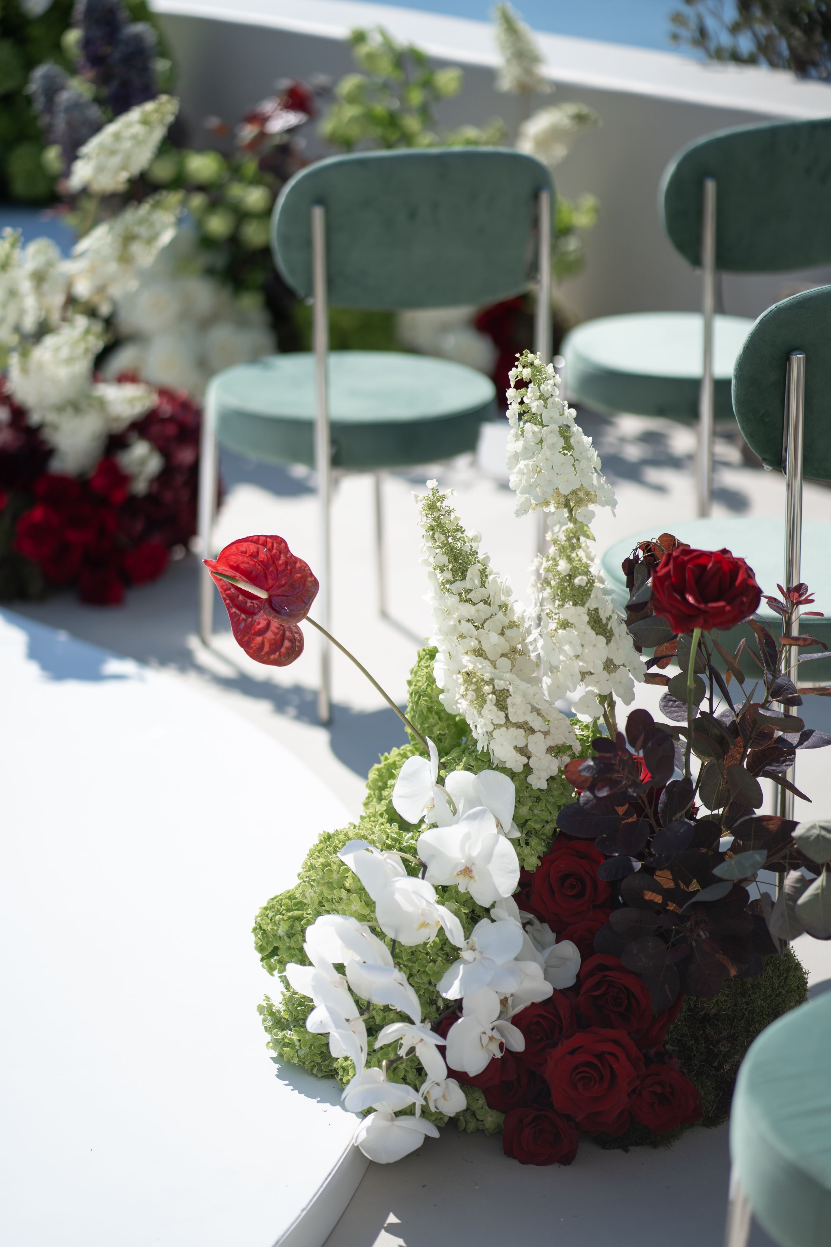 A close-up of a ceremony aisle arrangement on a sunlit white terrace at Islet in Santorini, Greece, set with wedding flowers. In the foreground, a tall spray of white hydrangea and green hydrangea sits beside a single red anthurium, white phalaenopsis orchids, a dark red rose and sprays of deep plum foliage. Behind it, pale green velvet chairs with slim silver legs line the aisle, with more white and red blooms softly blurred in the background.