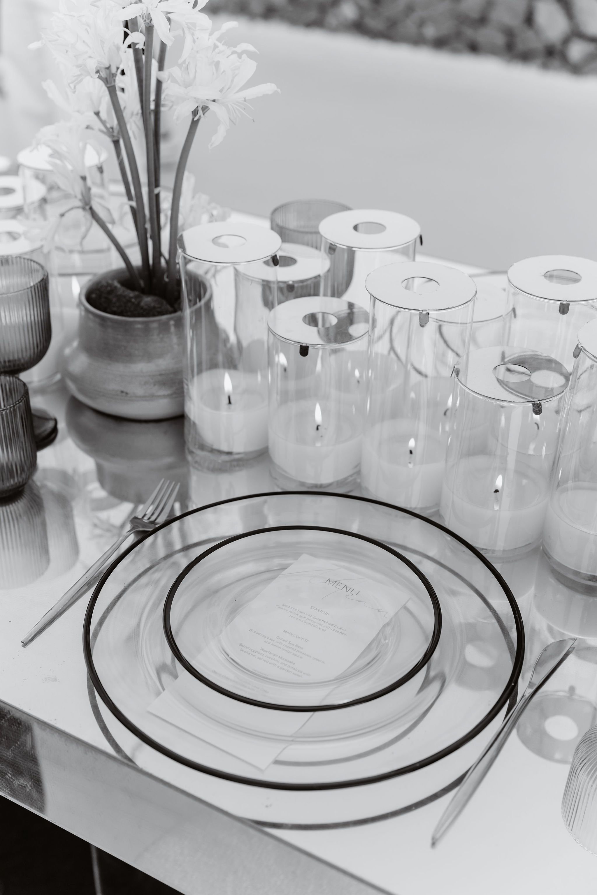 A black-and-white table setting at Aenaon Villas in Greece, styled for a wedding. Clear glass charger plates with dark rims hold a printed menu card, with silver cutlery on either side. Behind them stands a row of tall glass hurricane candle holders with lit white pillar candles, and a small ceramic pot with a dark green plant sits at the back left.