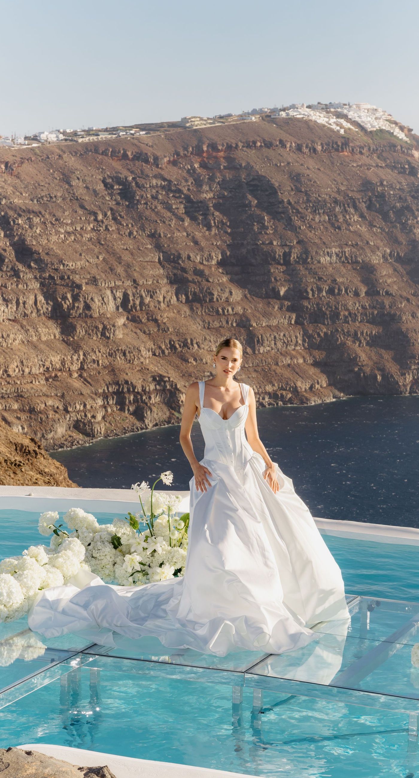 Emily & liam - In this exquisite scene, a couple stands in an ethereal embrace on a glass platform, surrounded by delicate blossoms that mirror the elegance of a sunlit seascape. The subdued pastels of the sunset wash gently over the horizon, casting a serene glow upon the couple and the blooms, creating a harmonious symphony of colors and emotions. The gracefully arranged white flowers echo timeless beauty and sophistication, set against the tranquil backdrop of a distant island. This stunning tableau invites you into a world where nature and romance converge seamlessly, an ideal representation for any florist's portfolio that values artistry and grace. - Photo 1