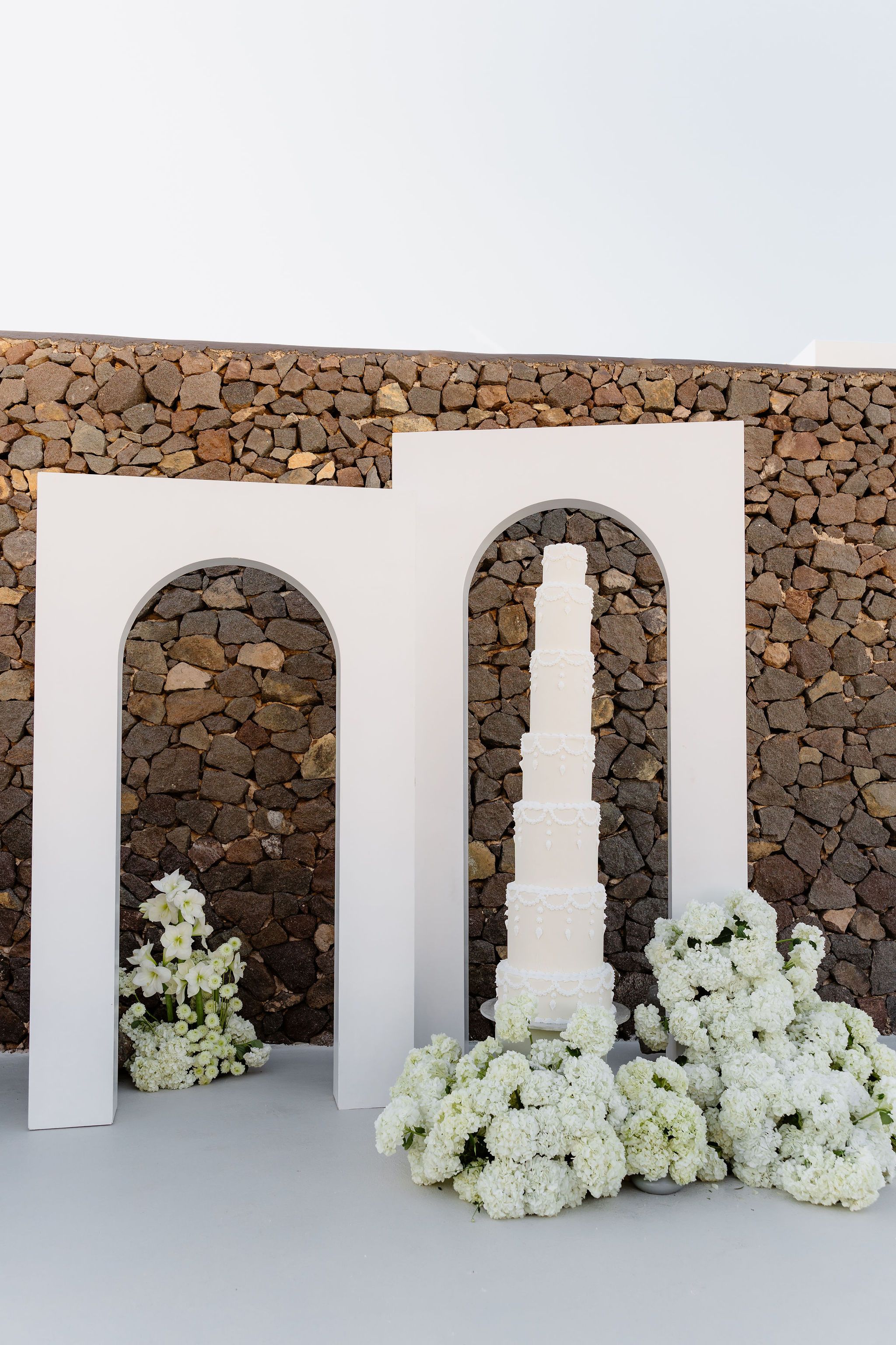 A tall, slender white wedding cake with piped scalloped garlands and drops stands before two white arches set against a dry-stone wall, with white flowers heaped at its base, all arranged as a wedding display at Aenaon Villas in Greece. A cluster of white stock and amaryllis blooms sits at the left arch, and the bright, soft light gives the scene a calm, minimal feel.