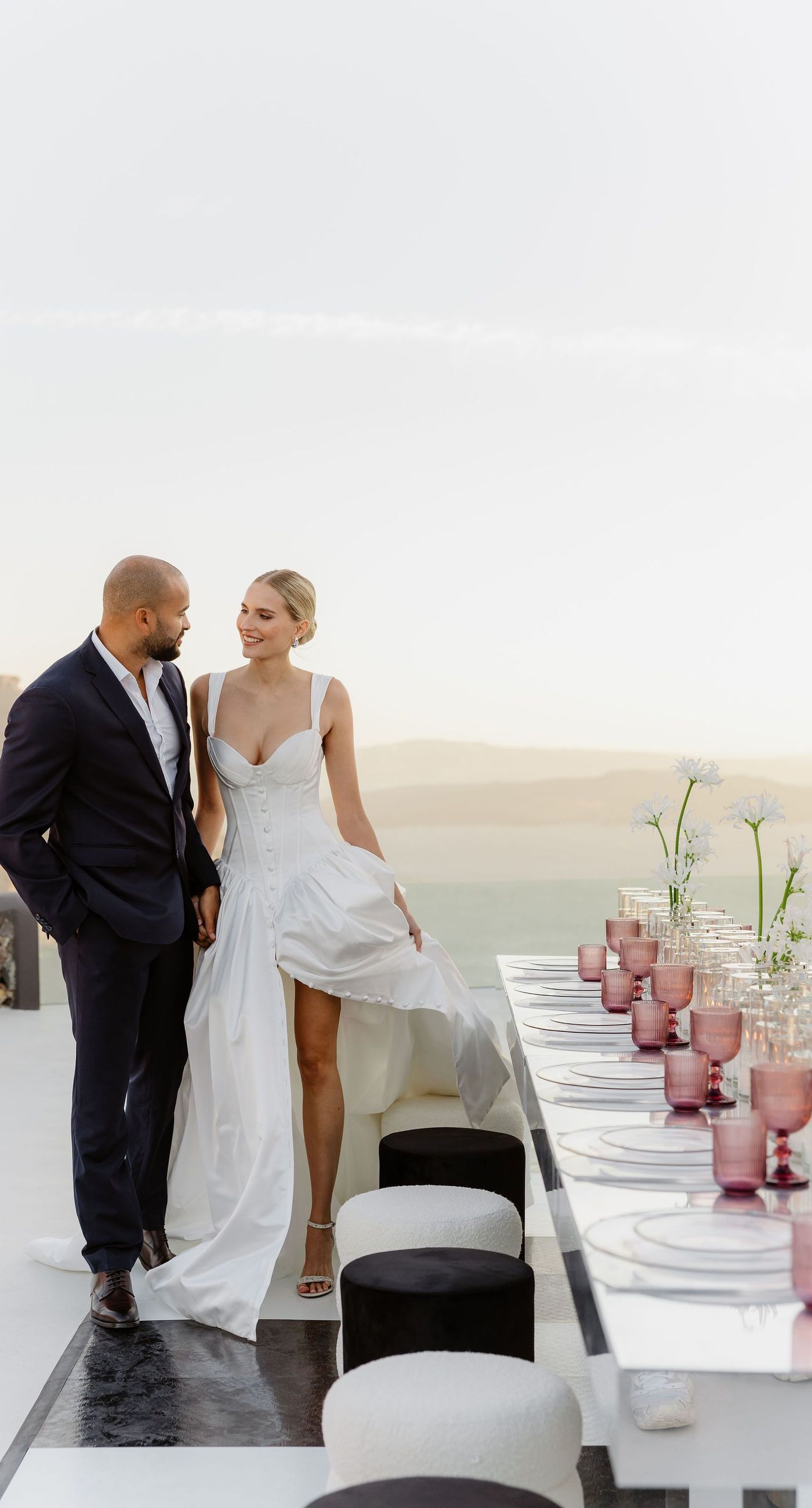 Emily & liam - In this exquisite scene, a couple stands in an ethereal embrace on a glass platform, surrounded by delicate blossoms that mirror the elegance of a sunlit seascape. The subdued pastels of the sunset wash gently over the horizon, casting a serene glow upon the couple and the blooms, creating a harmonious symphony of colors and emotions. The gracefully arranged white flowers echo timeless beauty and sophistication, set against the tranquil backdrop of a distant island. This stunning tableau invites you into a world where nature and romance converge seamlessly, an ideal representation for any florist's portfolio that values artistry and grace. - Photo 5