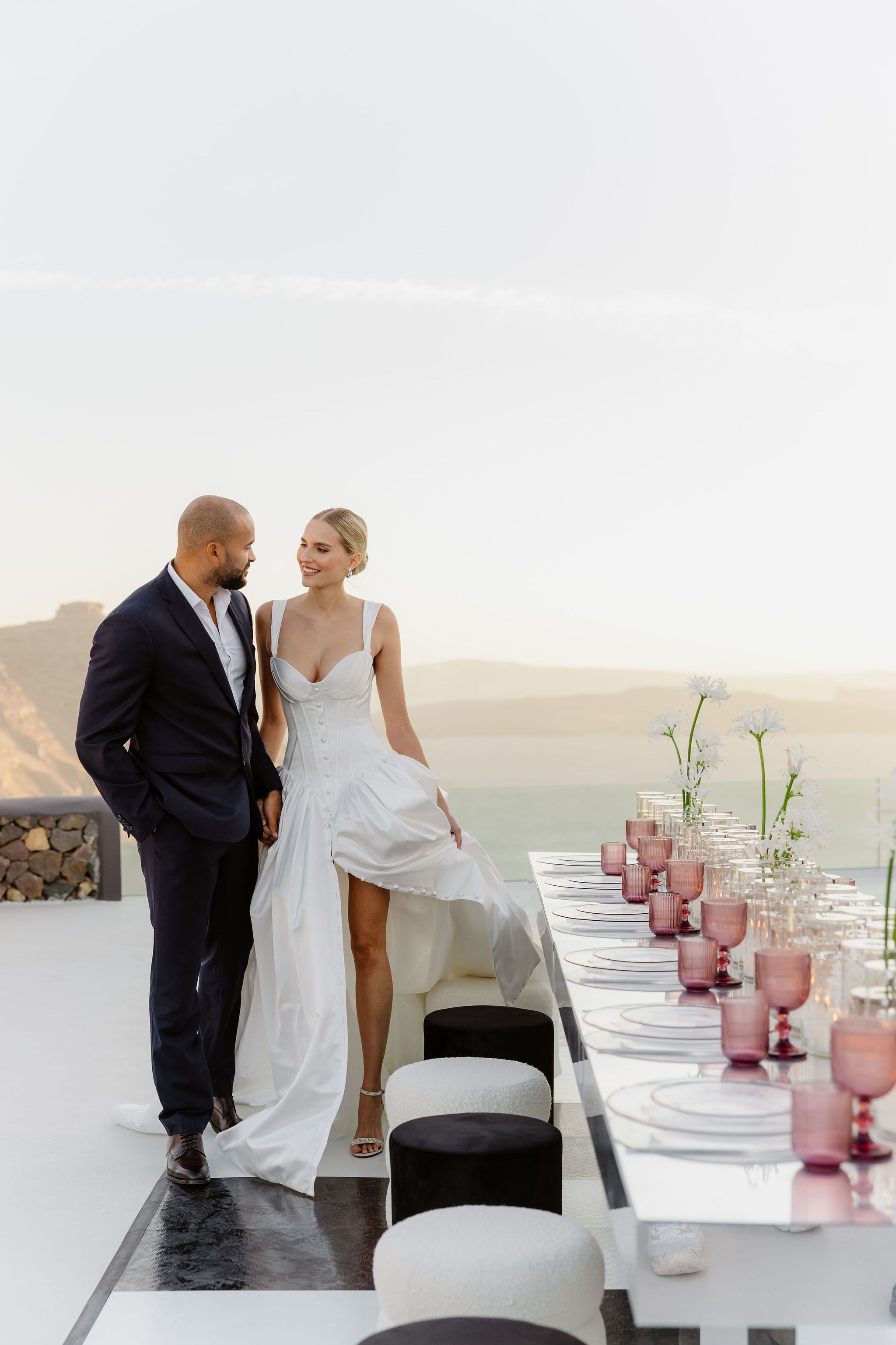 A bride in a white satin corset gown with a high slit, holding hands with a groom in a navy suit, stands on a pale terrace beside a long white table set with clear charger plates, rose-pink goblets and slender white agapanthus stems. Wedding table flowers and styling at Aenaon Villas in Greece, with a soft hazy sea and hillside behind them in golden evening light.
