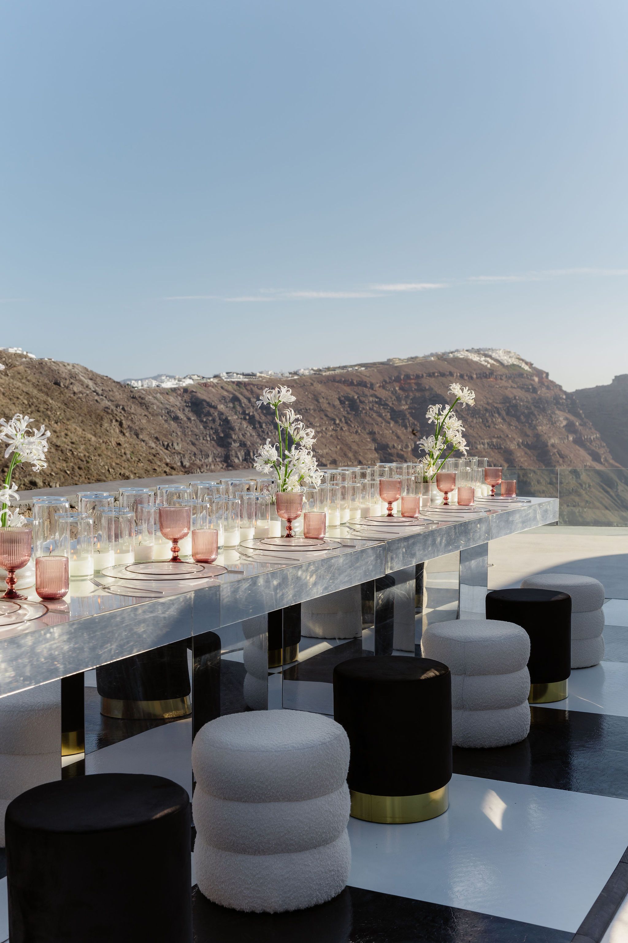 A long mirrored wedding table on a terrace at Aenaon Villas in Santorini, Greece, set with clear glass plates, blush pink stemmed goblets and tall white candles in glass hurricanes. Three tall arrangements of white tuberose-style flowers rise from the table, and the cliffside landscape with whitewashed village rooftops glows in warm evening light behind it. Round stools in cream bouclé and black velvet with brass bases sit along the checkered black-and-white floor.