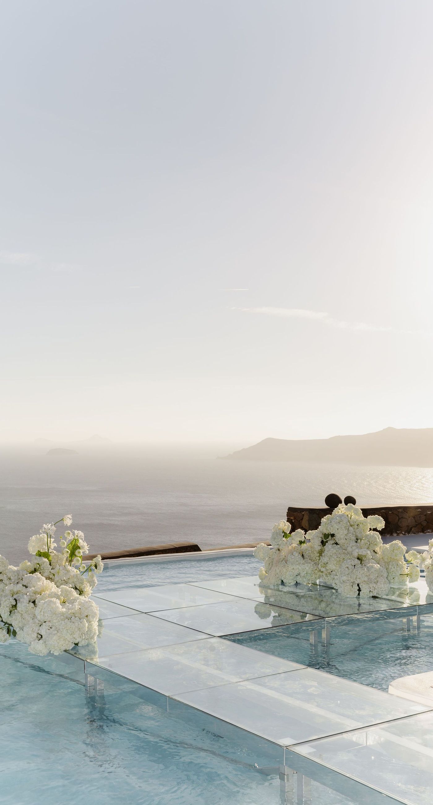 Emily & liam - In this exquisite scene, a couple stands in an ethereal embrace on a glass platform, surrounded by delicate blossoms that mirror the elegance of a sunlit seascape. The subdued pastels of the sunset wash gently over the horizon, casting a serene glow upon the couple and the blooms, creating a harmonious symphony of colors and emotions. The gracefully arranged white flowers echo timeless beauty and sophistication, set against the tranquil backdrop of a distant island. This stunning tableau invites you into a world where nature and romance converge seamlessly, an ideal representation for any florist's portfolio that values artistry and grace. - Photo 2