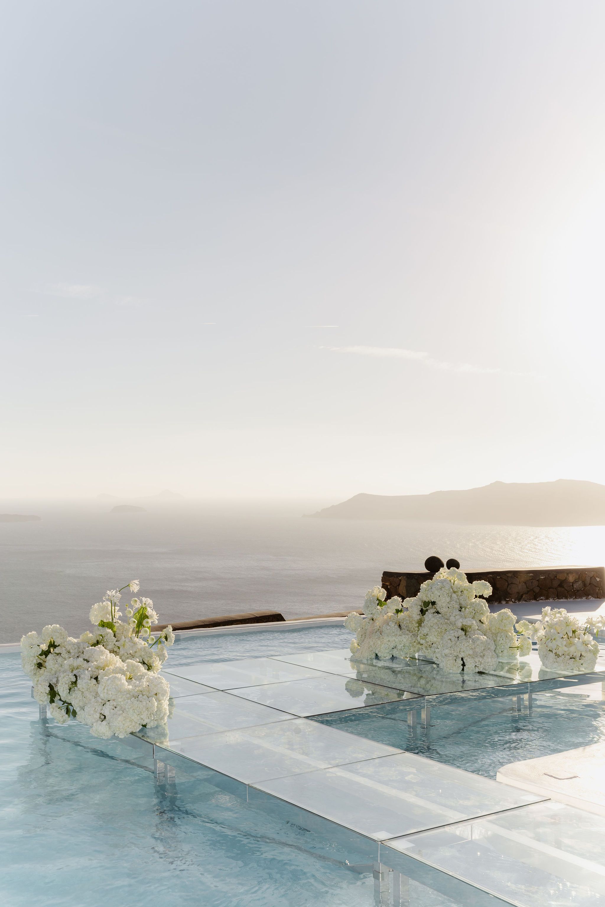 Clusters of white hydrangea and white blooms, with trailing green stems, rest on a glass walkway laid across an infinity pool at Aenaon Villas, a wedding flower setup in Greece. Pale turquoise water shows beneath the clear panels, and beyond the pool's edge the calm sea stretches toward hazy islands under a soft, sunlit sky. The overall mood is airy, serene and luminous.
