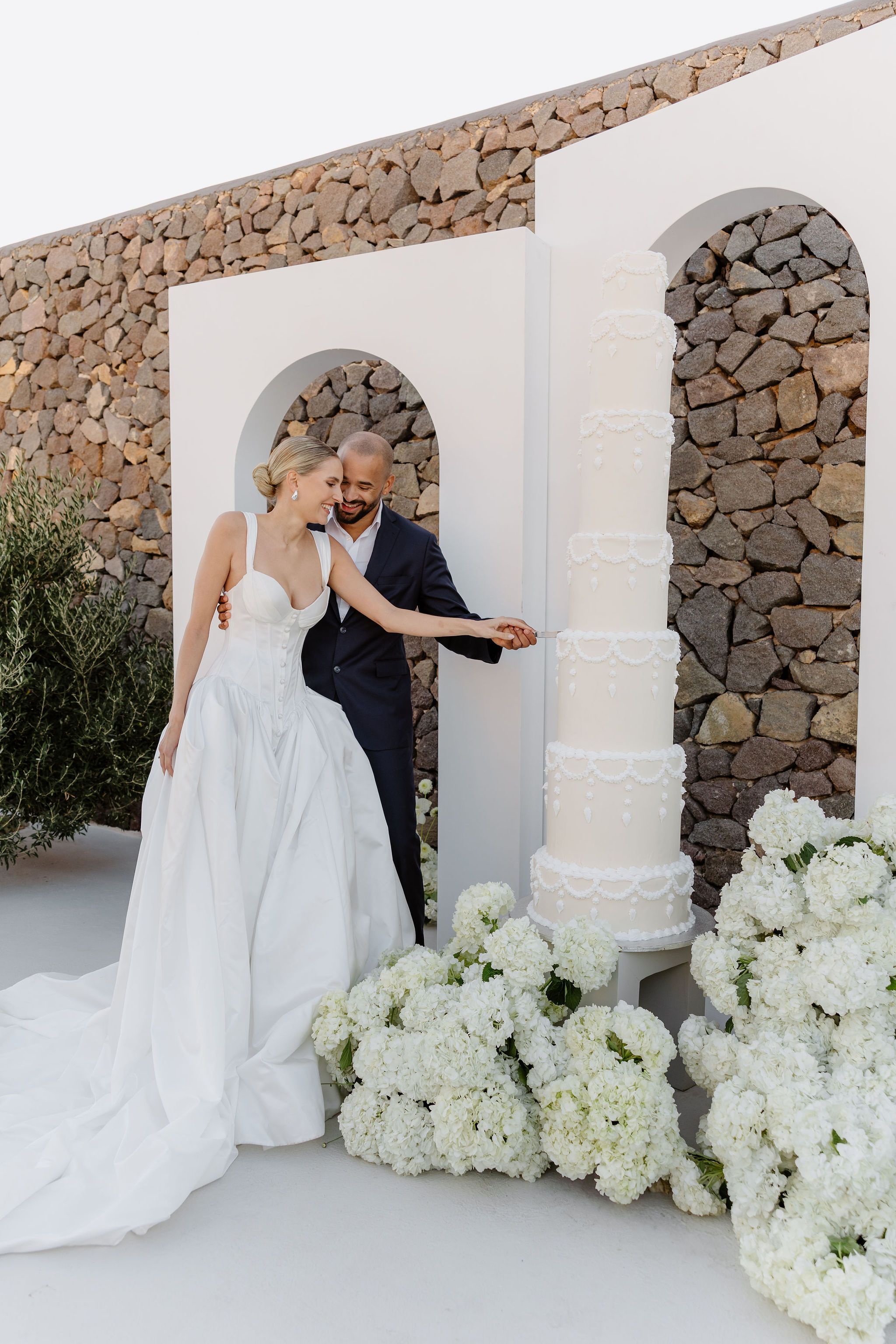 A bride in a white structured gown with a corset bodice and full taupe-white skirt laughs as she and her groom, in a navy suit with an open white collar, guide a knife through a tall, five-tier white cake decorated with piped scallops. Clusters of white hydrangeas fill the foreground, and an arched white niche sits against a dry-stone wall at Aenaon Villas, a wedding venue in Greece, with olive foliage at the left.