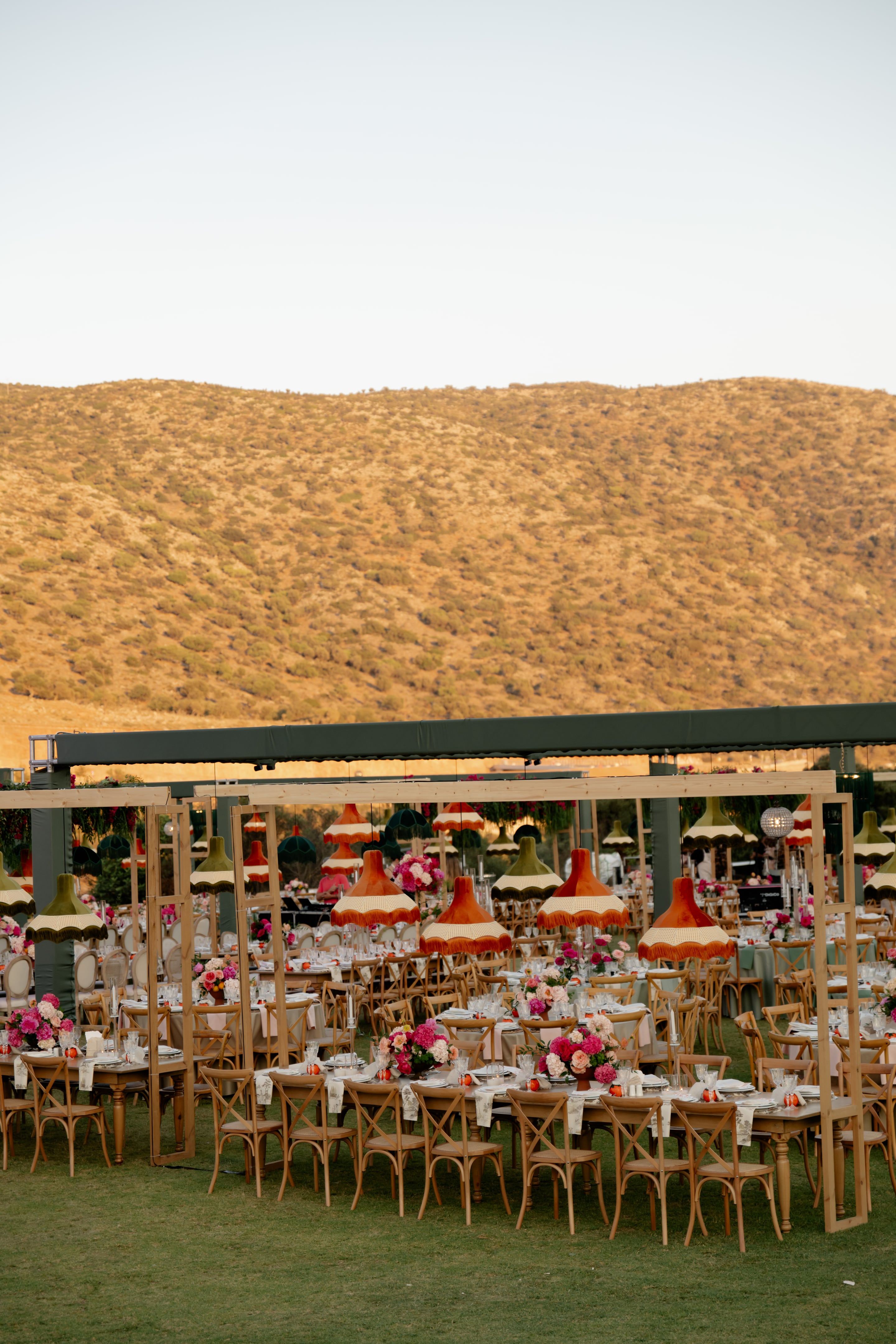 Long wooden trestle tables with cross-back chairs fill an open-sided marquee on a lawn, set for a wedding dinner in Crete, Greece. Pink and white peonies and roses sit in low centrepieces, and rows of fringed lampshades in orange, olive green and cream hang above, while golden, sunlit hills rise behind the tent in the evening light.