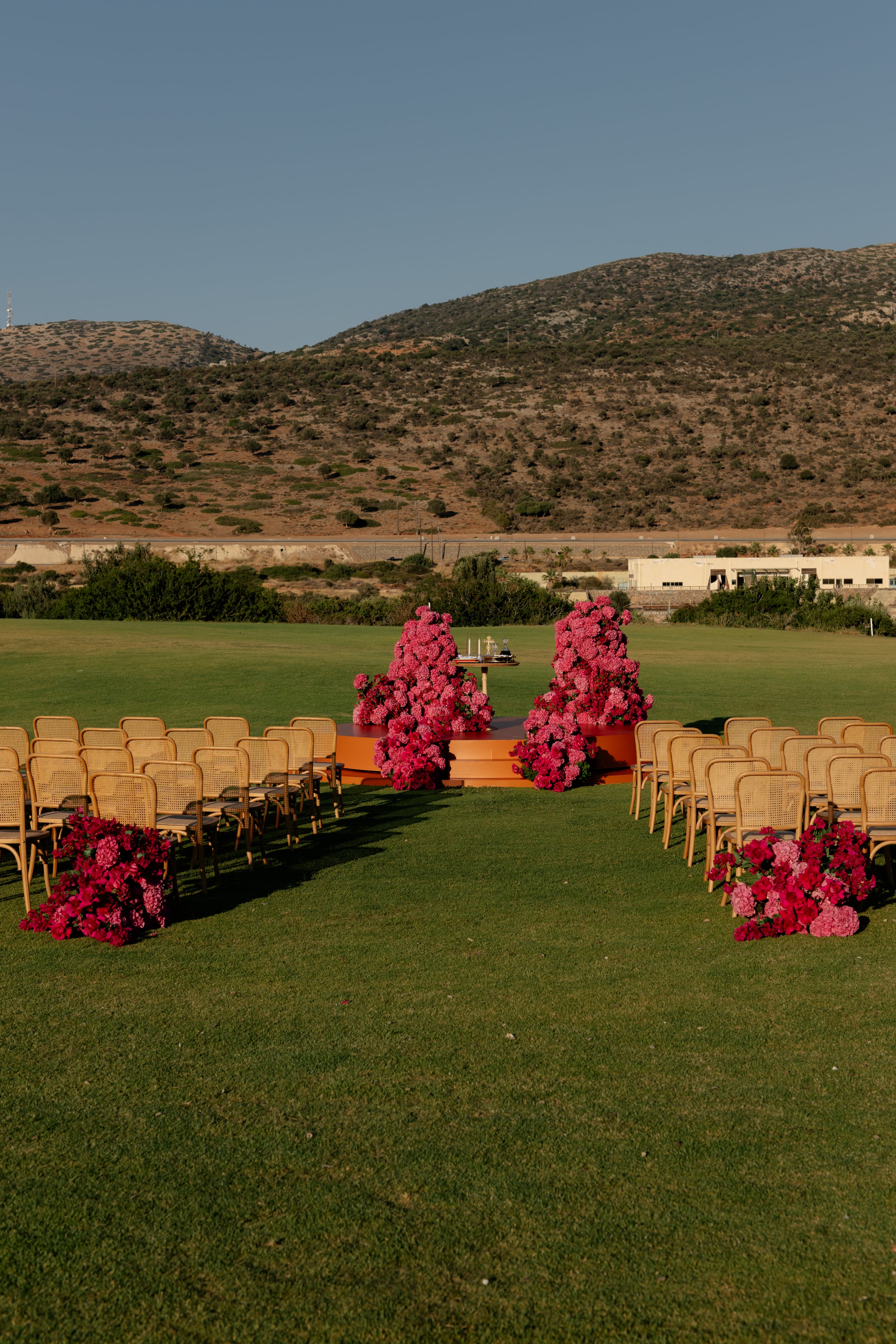 Two tall, sculptural mounds of pink and magenta hydrangeas and bougainvillea rise from a low orange platform at the far end of a grassy lawn, with a small gold table holding candles between them. Rows of woven cane chairs line both sides of an aisle that is edged with cascading clusters of red and pink blooms. Behind, dry Cretan hills and a pale building sit under a clear late-afternoon sky. This is a wedding ceremony set-up in Crete, Greece, at Crete Golf Club.