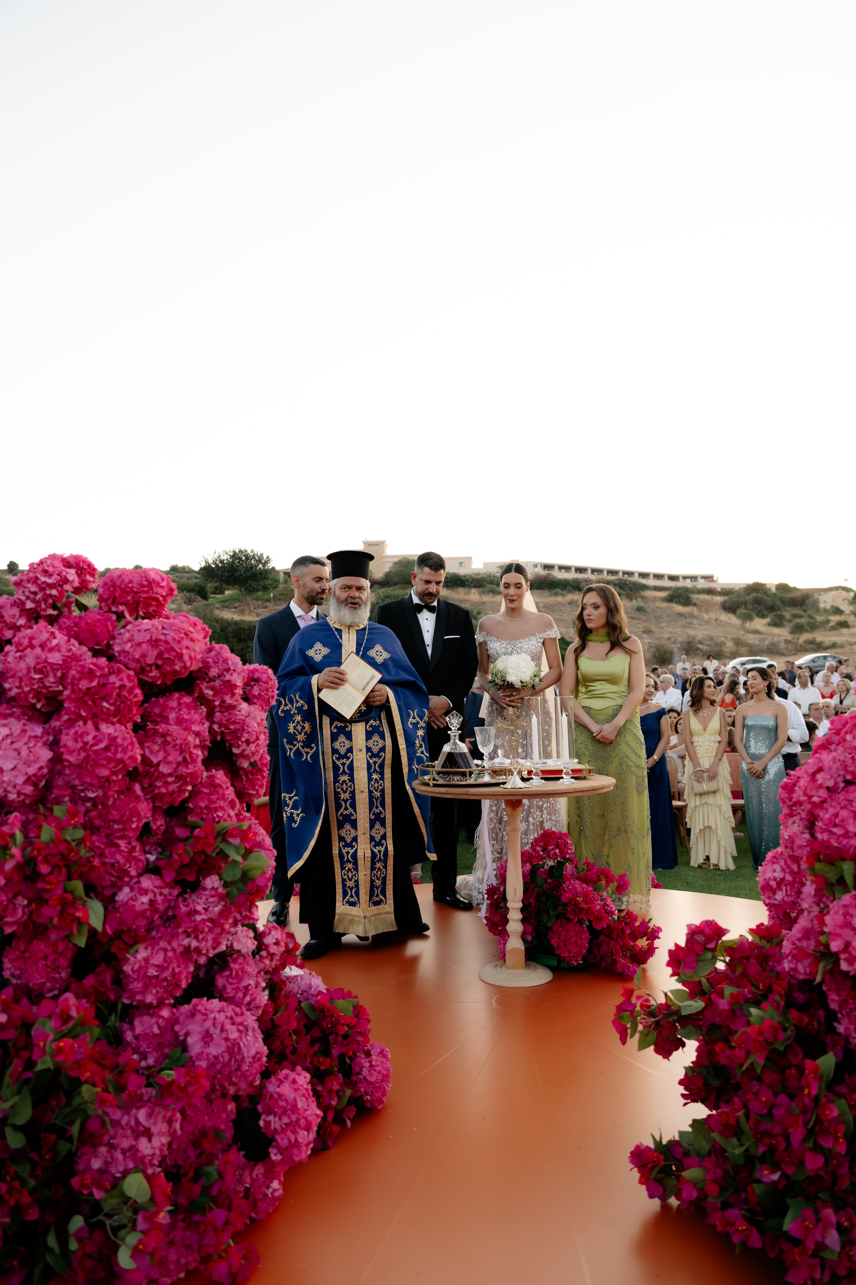 Pink hydrangeas and magenta bougainvillea frame the foreground of this outdoor wedding ceremony at Crete Golf Club in Crete, Greece, with the flowers rising in dense, sculptural clusters on either side of an orange-red floor. At the centre, an Orthodox priest in a royal blue and gold vestment holds a prayer book, flanked by a groom in a black tuxedo and a bride in a sparkling, beaded gown holding a white bouquet. A bridesmaid in a sage-green satin gown stands beside a small wooden table set with glasses and candles, and guests in soft summer tones watch on a sunlit hillside.
