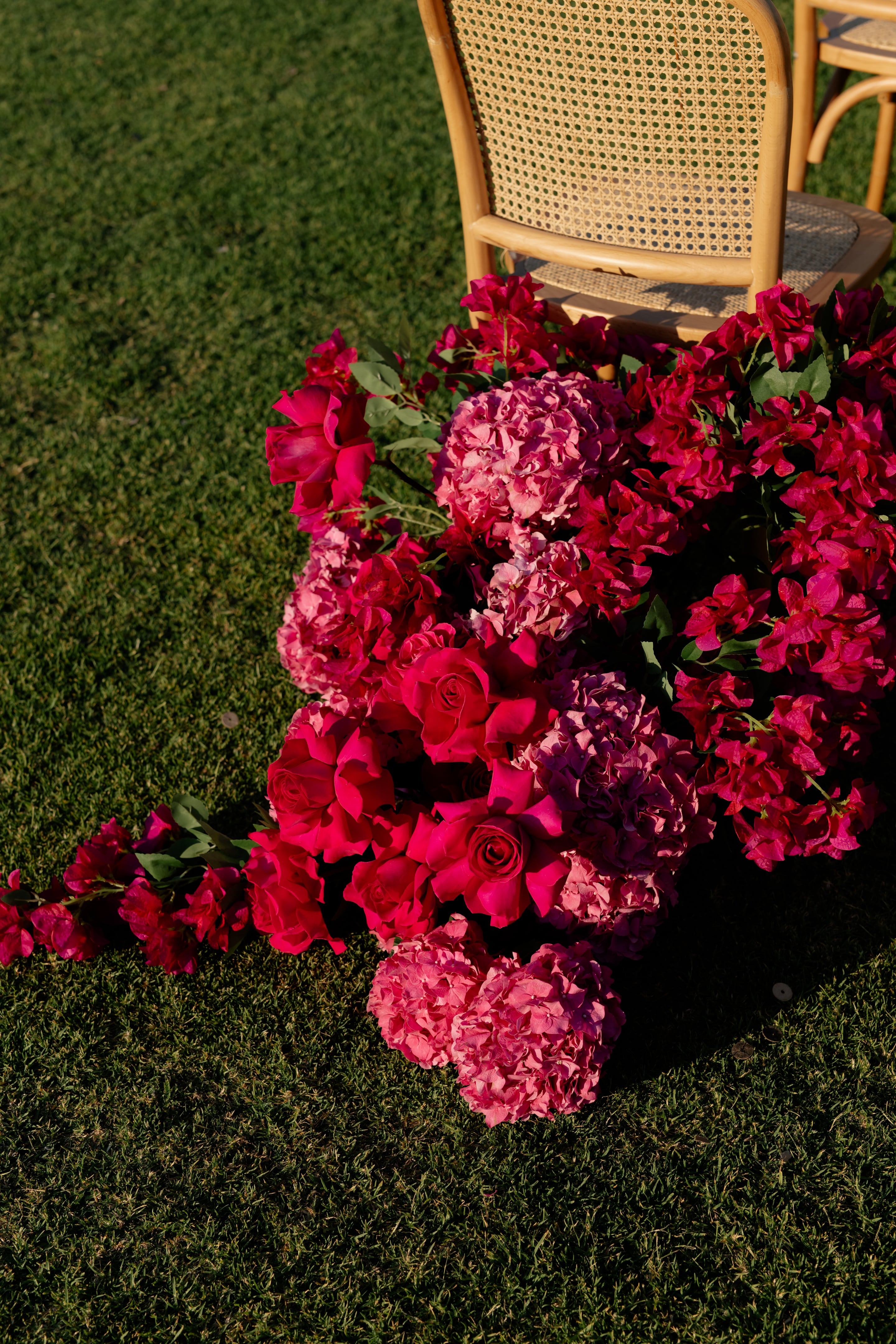 A cascading mound of hot pink and magenta flowers spills across green lawn at the foot of a woven cane chair, part of a wedding ceremony set up at Crete Golf Club in Greece. Deep pink roses sit among soft pink and mauve hydrangea heads and clusters of bougainvillea, with dark green leaves threading through the arrangement. Warm late-afternoon sunlight falls across the grass and the rich, saturated blooms, giving the scene a vivid, romantic feel.