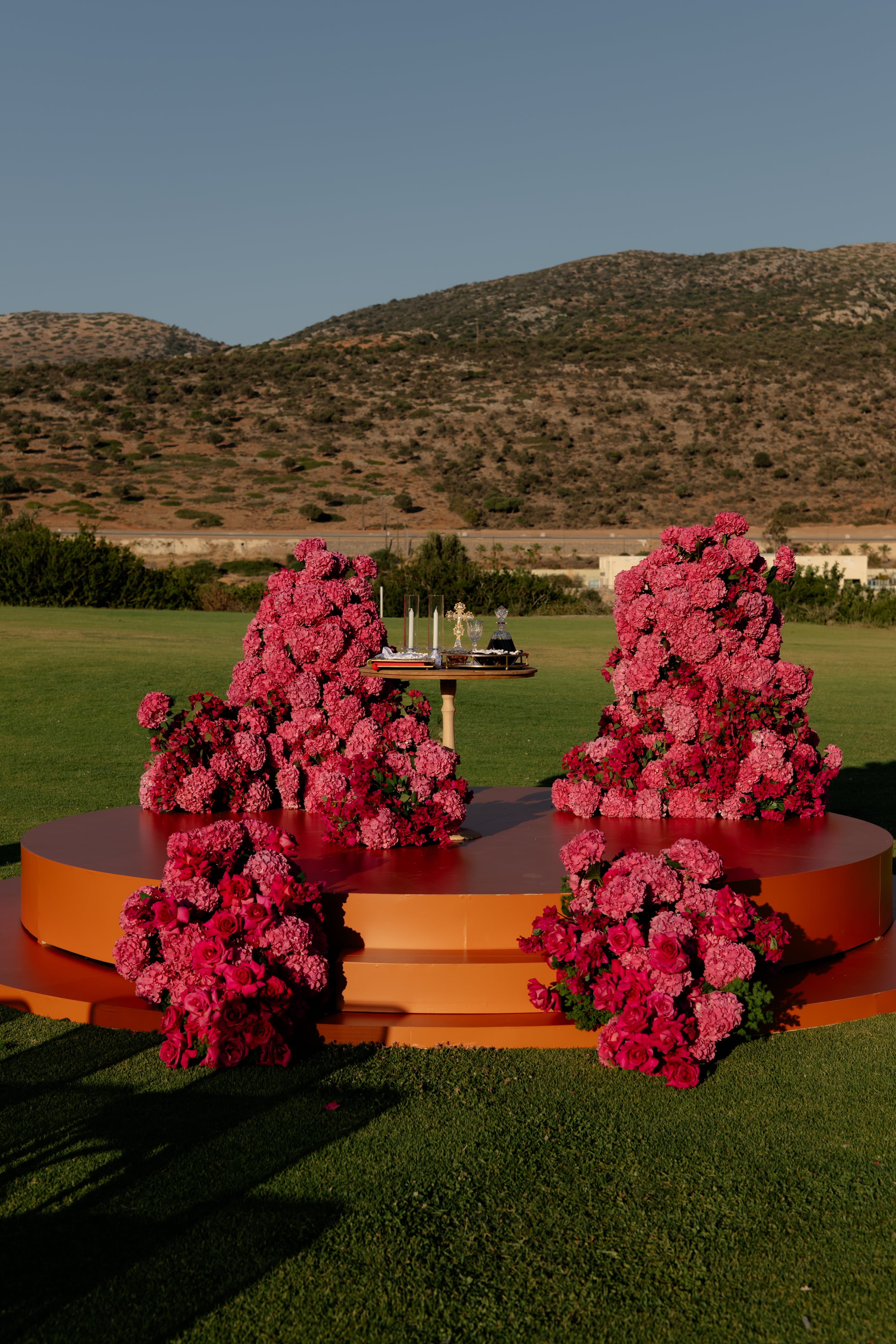 Two tall, cascading mounds of pink hydrangeas and deep rose-red blooms flank a small gold-legged table holding a cross, candles and a glass carafe, all set on a tiered orange stage on a green lawn at Crete Golf Club in Crete, Greece. Smaller low clusters of hot pink roses and pale pink hydrangeas sit at the stage's front corners, and in the warm late-afternoon light, the whole wedding ceremony setup glows against the dry brown hills behind.