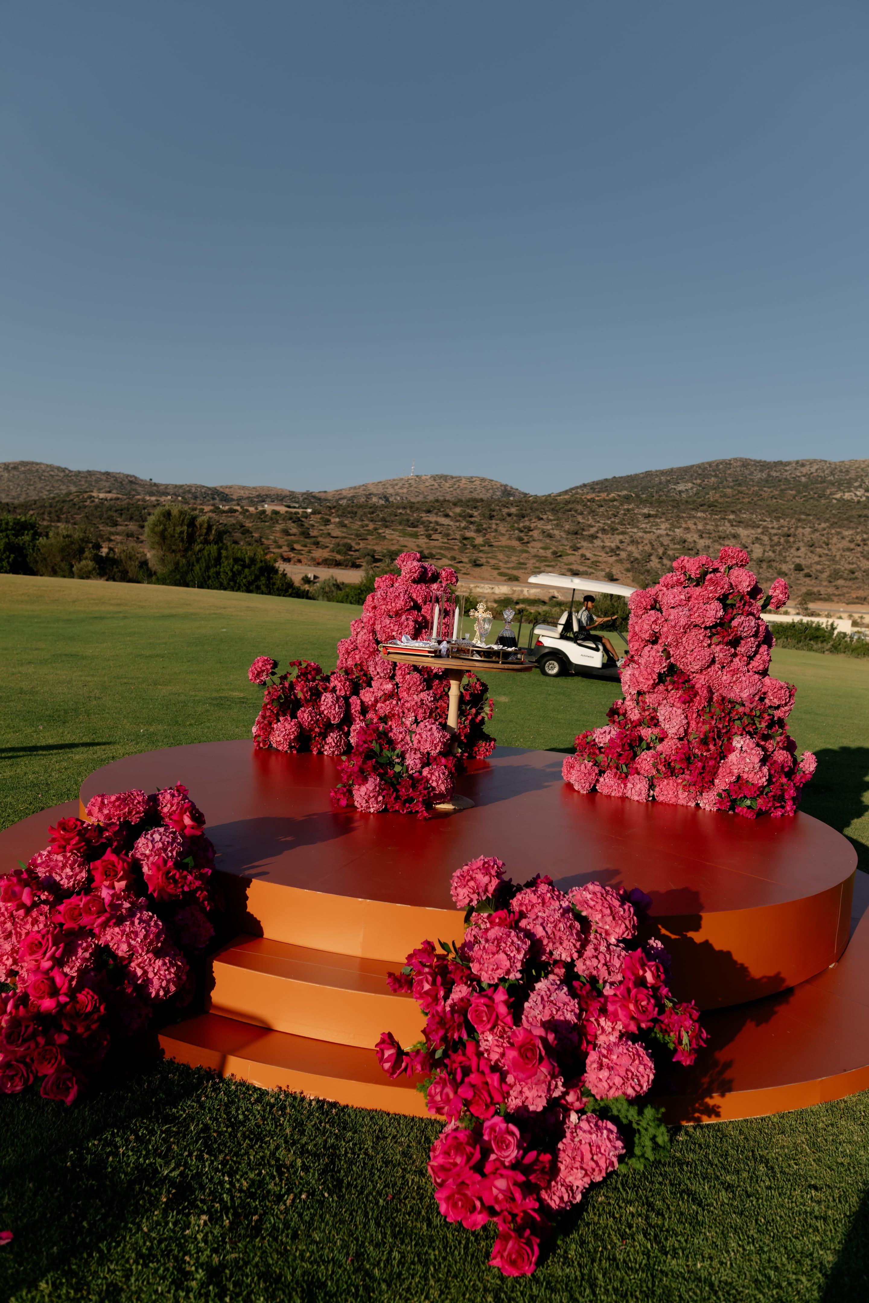 Tiered terracotta-orange circular platforms on a green lawn hold dense clusters of pink hydrangeas and deep pink garden roses, with a small table at the centre set with candles and silver chalices. A golf cart passes in the sunlit background above the grass. This wedding ceremony set-up, with its sculptural pink floral installations, is at Crete Golf Club in Crete, Greece.
