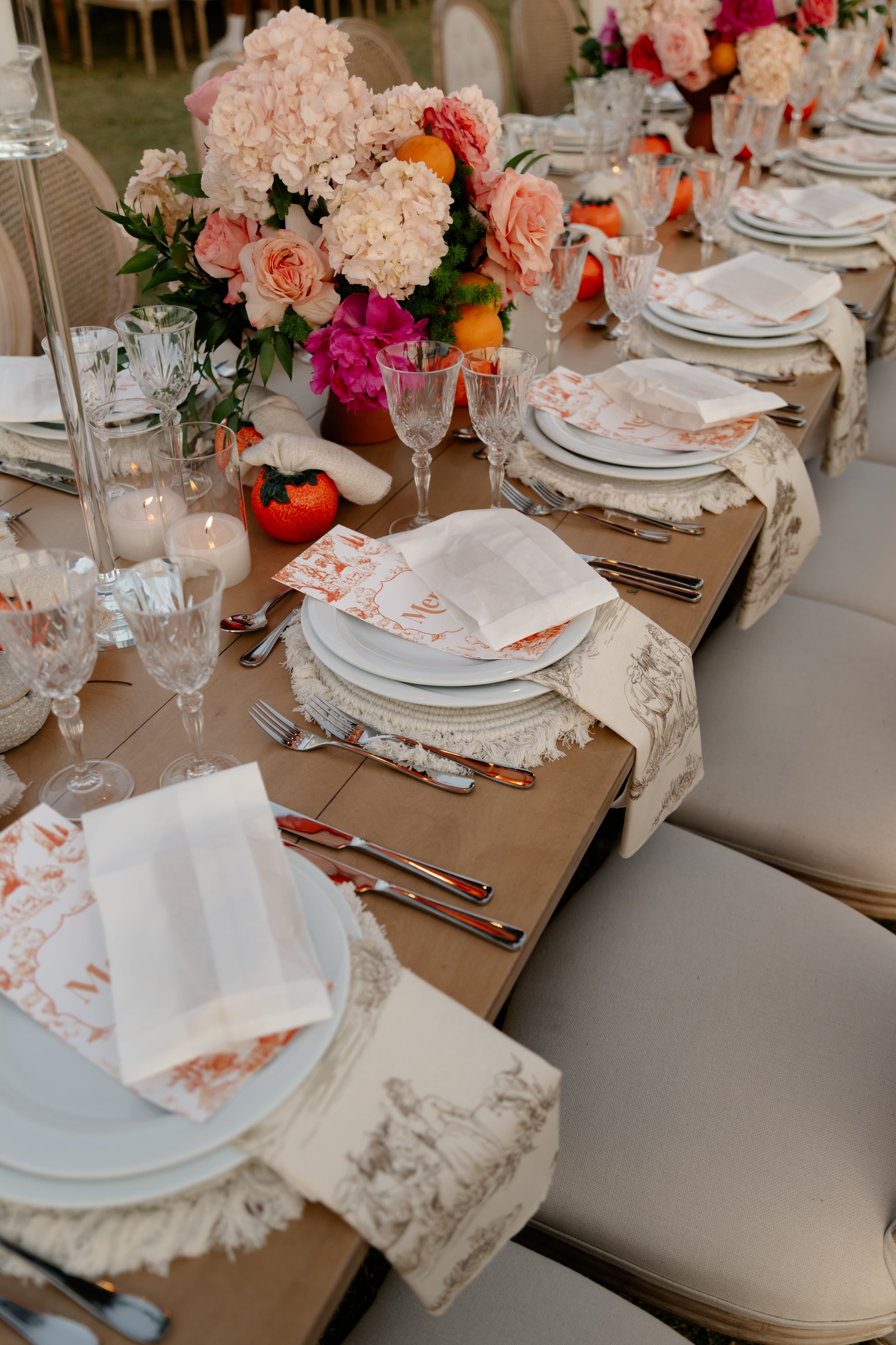 A long wooden wedding table laid for a celebration in Greece, with a centrepiece of cream hydrangeas, blush garden roses and a hot pink peony rising from a terracotta vase, tucked among oranges and sprigs of green foliage. Place settings hold white plates on fringed woven mats, crystal wine glasses, and cream napkins printed with rust-coloured toile, with orange-toned name cards. Glowing pillar candles and small orange fruits add warmth to the soft, evening-lit scene at Crete Golf Club, Crete, Greece.