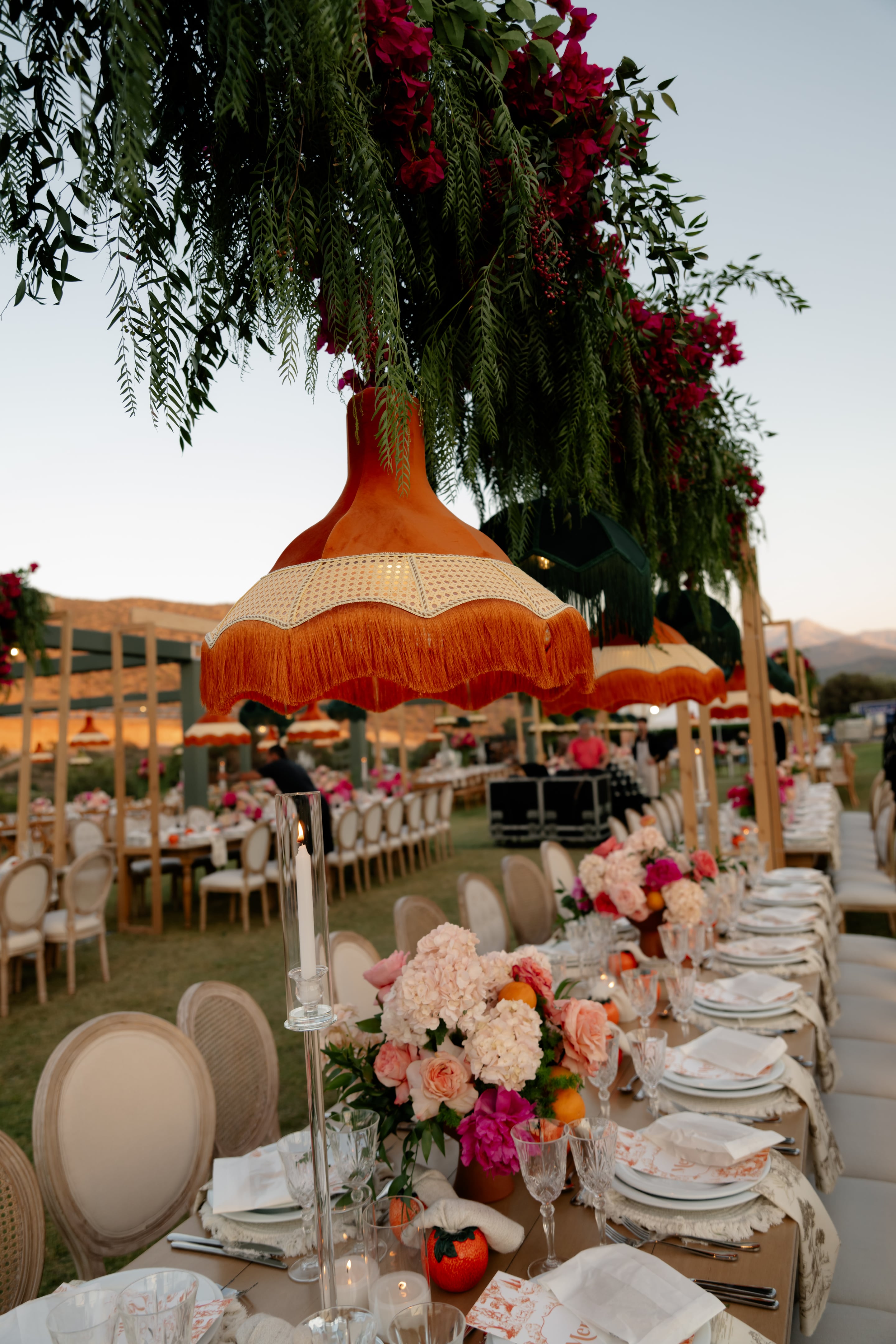 Long banquet tables stretch across a lawn at dusk, set with white plates, clear glassware and blush hydrangeas, garden roses and orange fruit in the centrepieces. Overhead hang terracotta lampshades with cane bands and orange fringe, beneath trailing green foliage and magenta bougainvillea. This wedding table flower styling is at cretegolfclub in Crete, Greece, with a candle in a glass hurricane holder in the foreground and mountains softly lit behind.