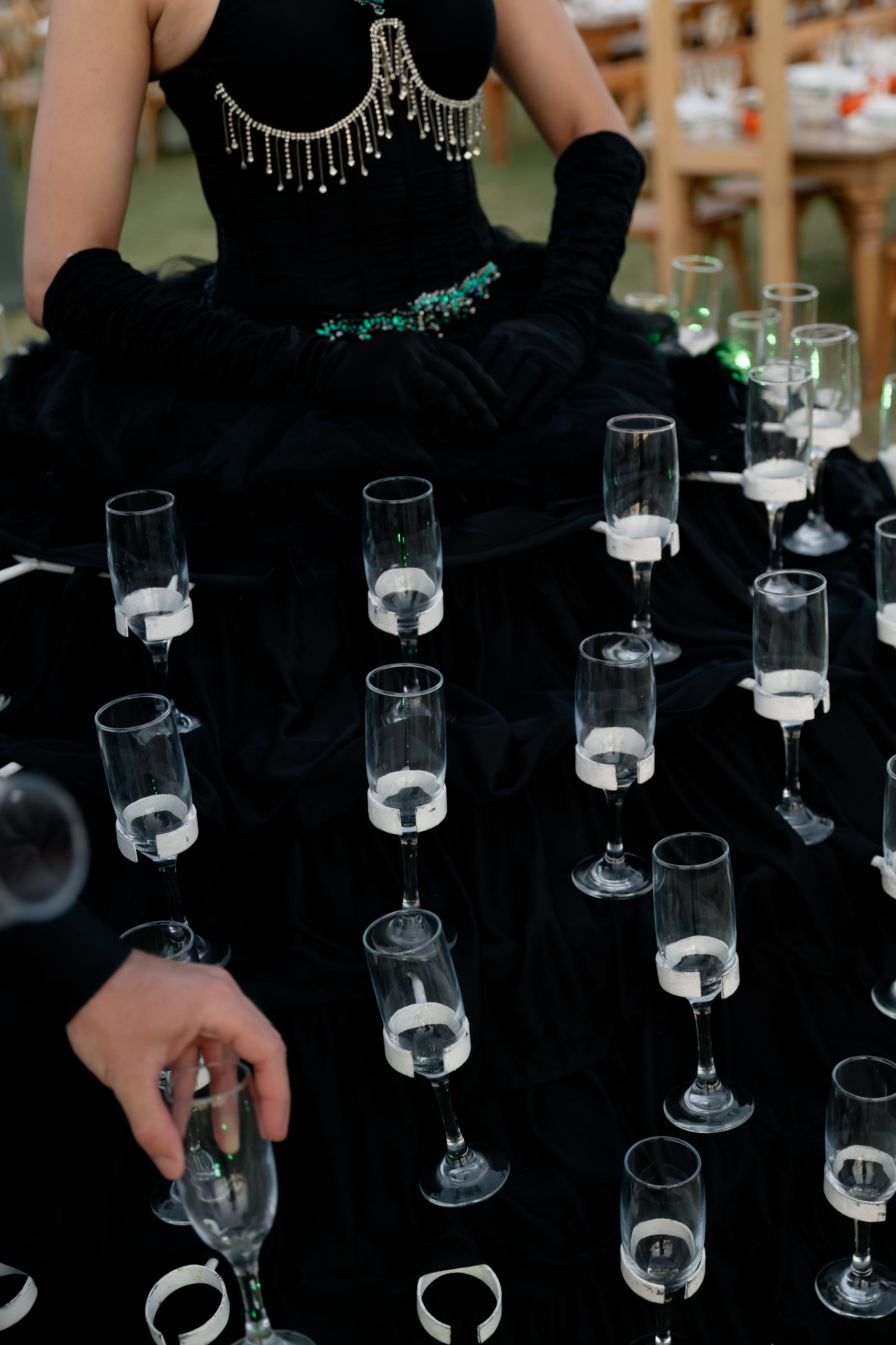 Rows of empty champagne flutes stand upright on a black fabric surface, each held in a white ring, with a figure in a black gown and long black gloves and a green beaded waistband seated behind them. The glasses catch soft outdoor light and reflect in the dark cloth. This is a sculptural installation for a wedding ceremony at Crete Golf Club in Crete, Greece, arranged as an unusual setting for the event.