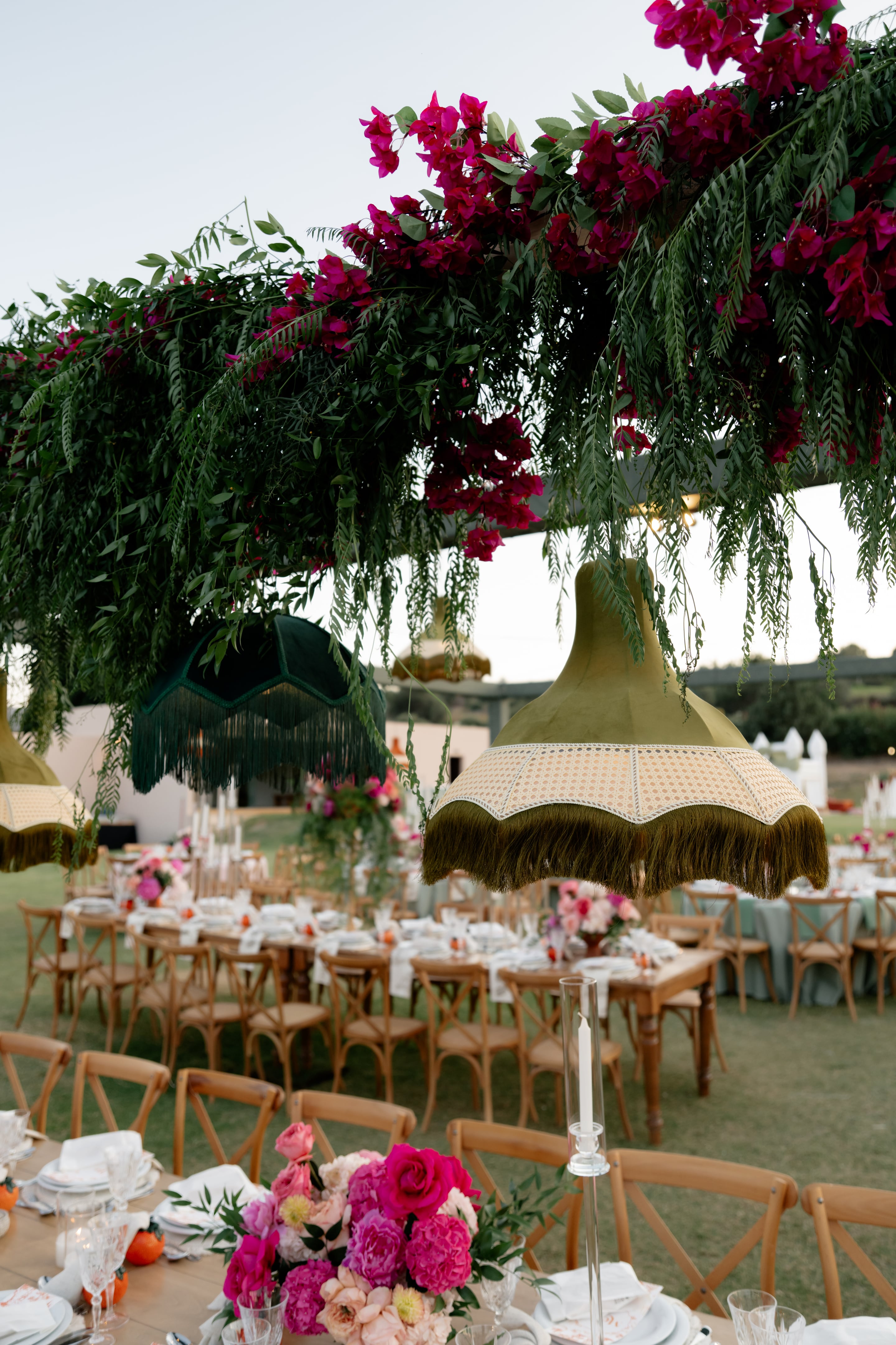 Suspended above long wooden banquet tables on a grassy lawn, oversized velvet lampshades in olive green and deep emerald hang beneath a canopy of trailing fern-like greenery and magenta bougainvillea, with woven cane bands and long fringe. Below, cross-back chairs surround tables set with white linens and small arrangements of pink roses and blooms, all part of a wedding flower and styling display in Crete, Greece, at the Crete Golf Club.