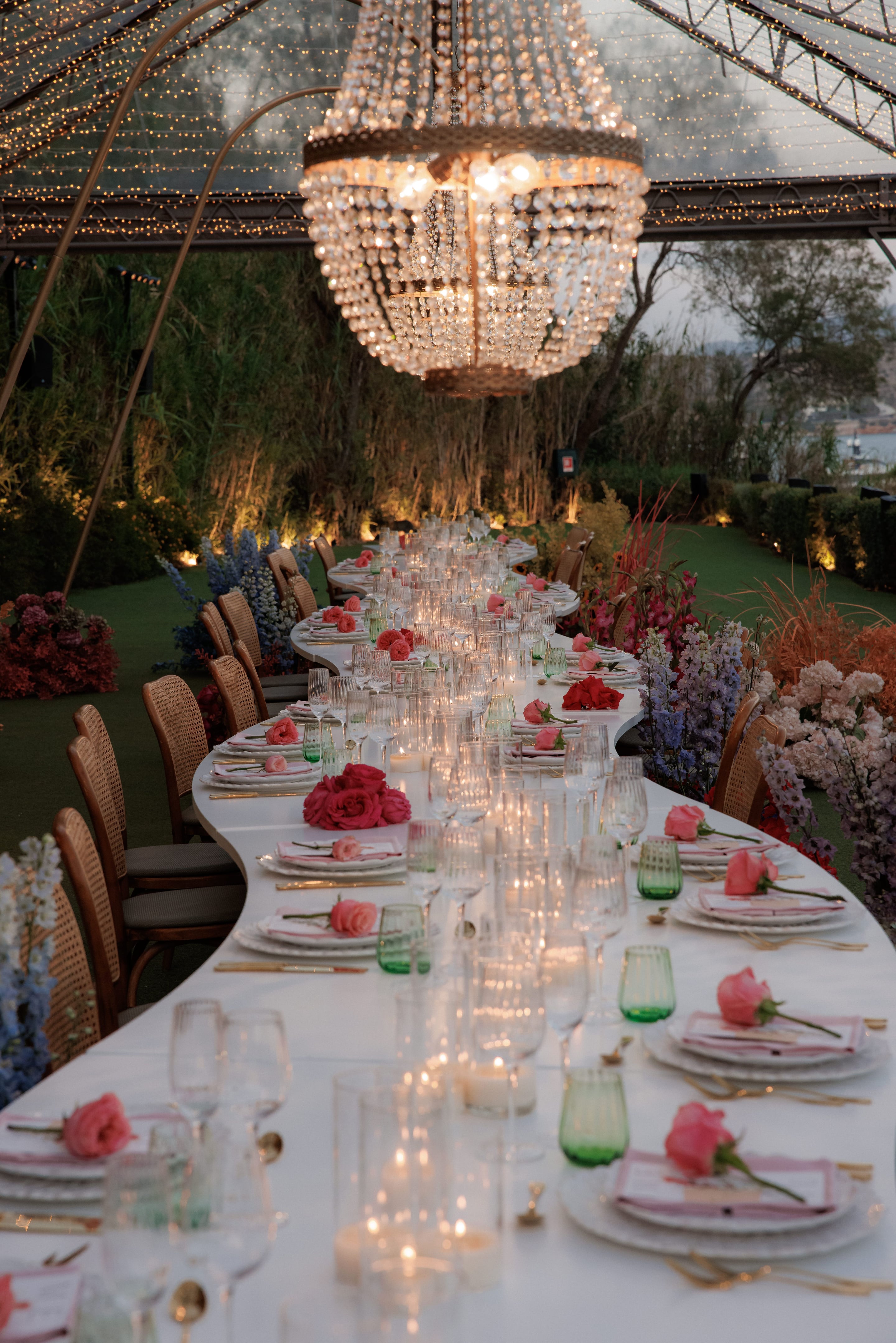 A long, curving white banquet table laid for a wedding dinner in Greece, set beneath a large crystal chandelier and strings of warm fairy lights under a clear canopy. Along the table sit clusters of deep pink garden roses, single pink roses on the plates, and tall glasses and candles, while tall blue and lavender delphinium and pale pink stock spill from beds of flowers at the sides. Rattan chairs line the table in the soft dusk light, with greenery and a glimpse of the sea beyond.
