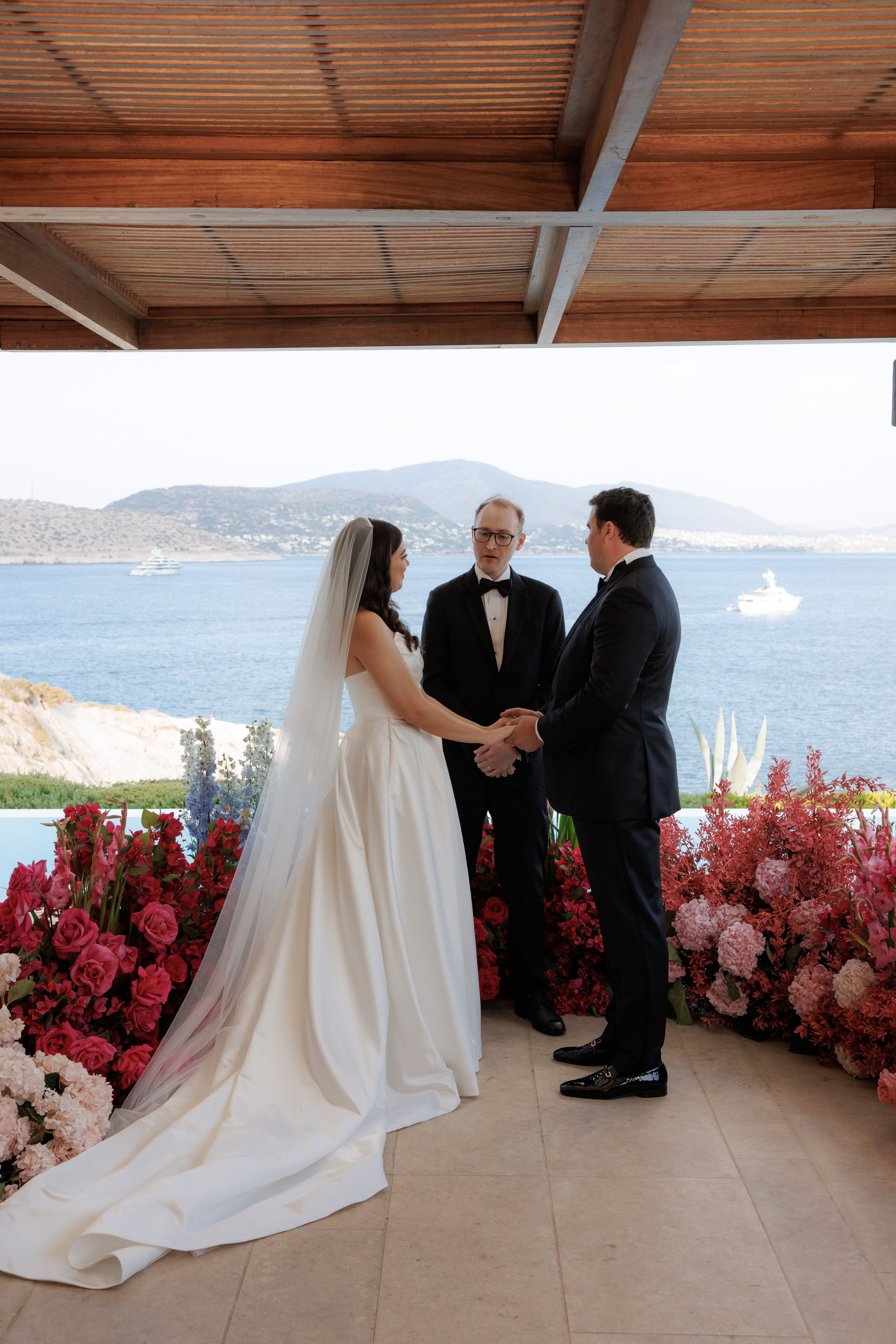 A wedding ceremony under a wooden pergola at Island Athens Riviera in Greece, with a bride in a strapless satin gown and long veil holding hands with her groom in a black tuxedo while an officiant in glasses and a bow tie stands between them. Below them, a low border of pink and red roses, carnations, and red spray flowers frames the aisle, with pale blue delphinium rising at the left. Behind them the sea glitters blue toward a hazy coastline, with two white yachts on the water.