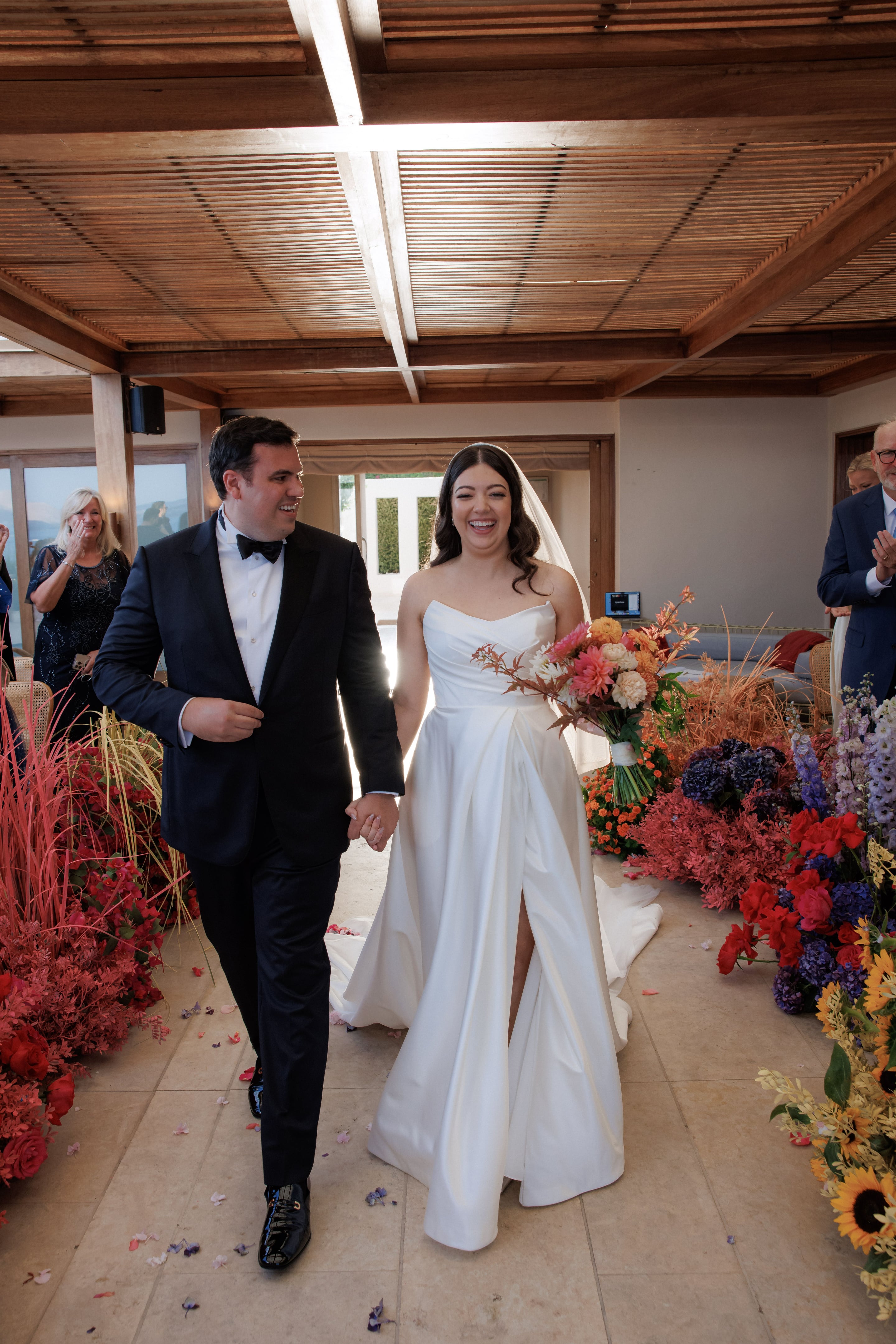 A bride in a strapless white gown and sheer veil laughs brightly as she walks hand in hand with a groom in a black tuxedo and bow tie, past guests applauding on either side of an aisle at Island Athens Riviera in Greece. The aisle is lined with a lush wedding floral installation of burnt orange and coral dahlias, cream blooms, purple hydrangeas, lavender delphinium and feathery russet grasses, all beneath a warm wooden ceiling.