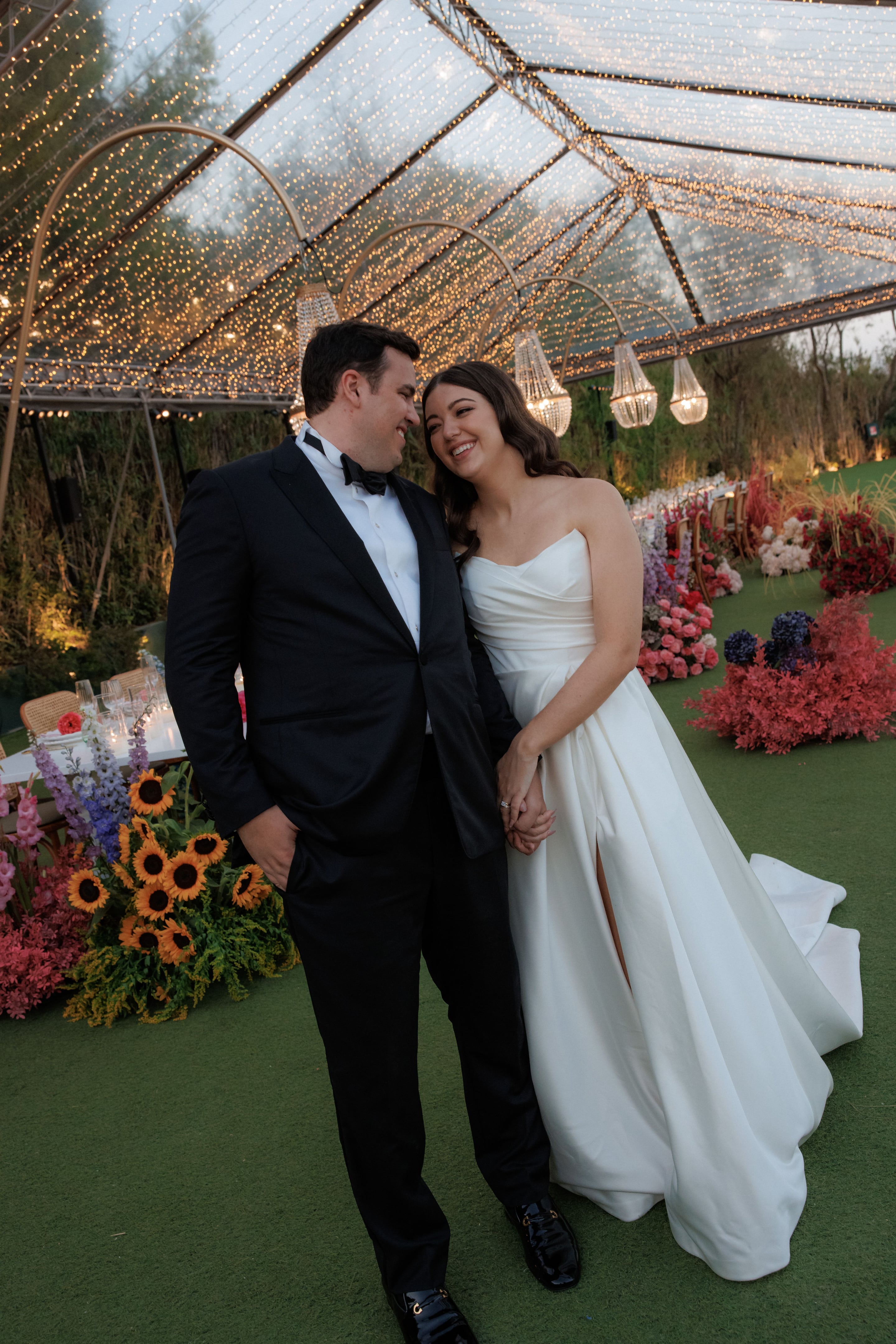 A bride in a strapless white satin gown with a thigh-high slit and a groom in a black tuxedo with bow tie stand close together, both laughing, beneath a glass-roofed tent strung with fairy lights and hung with crystal chandeliers. In the foreground are sunflowers and purple delphiniums, and beyond them a long reception table is lined with red, pink and white blooms, wedding table flowers at Island Athens Riviera in Greece.