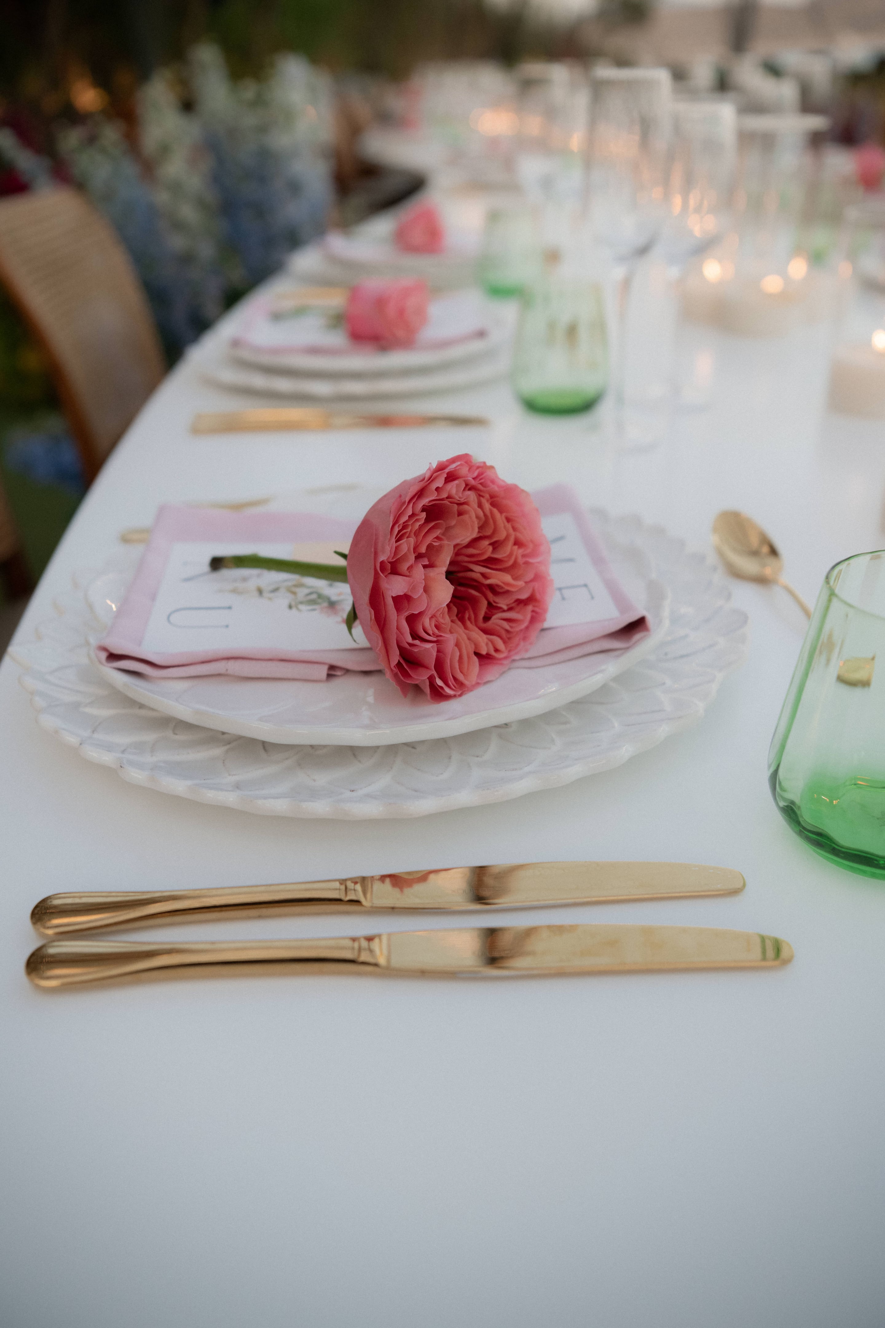 A long white wedding table set with a single coral-pink garden rose lying across a pale pink napkin and a scalloped white plate, with a gold spoon and green glass tumblers beside it. Soft candlelight glows in the background, and blurred blue and white blooms line the far end of the table. The flower is for a wedding table at Island Athens Riviera in Greece.