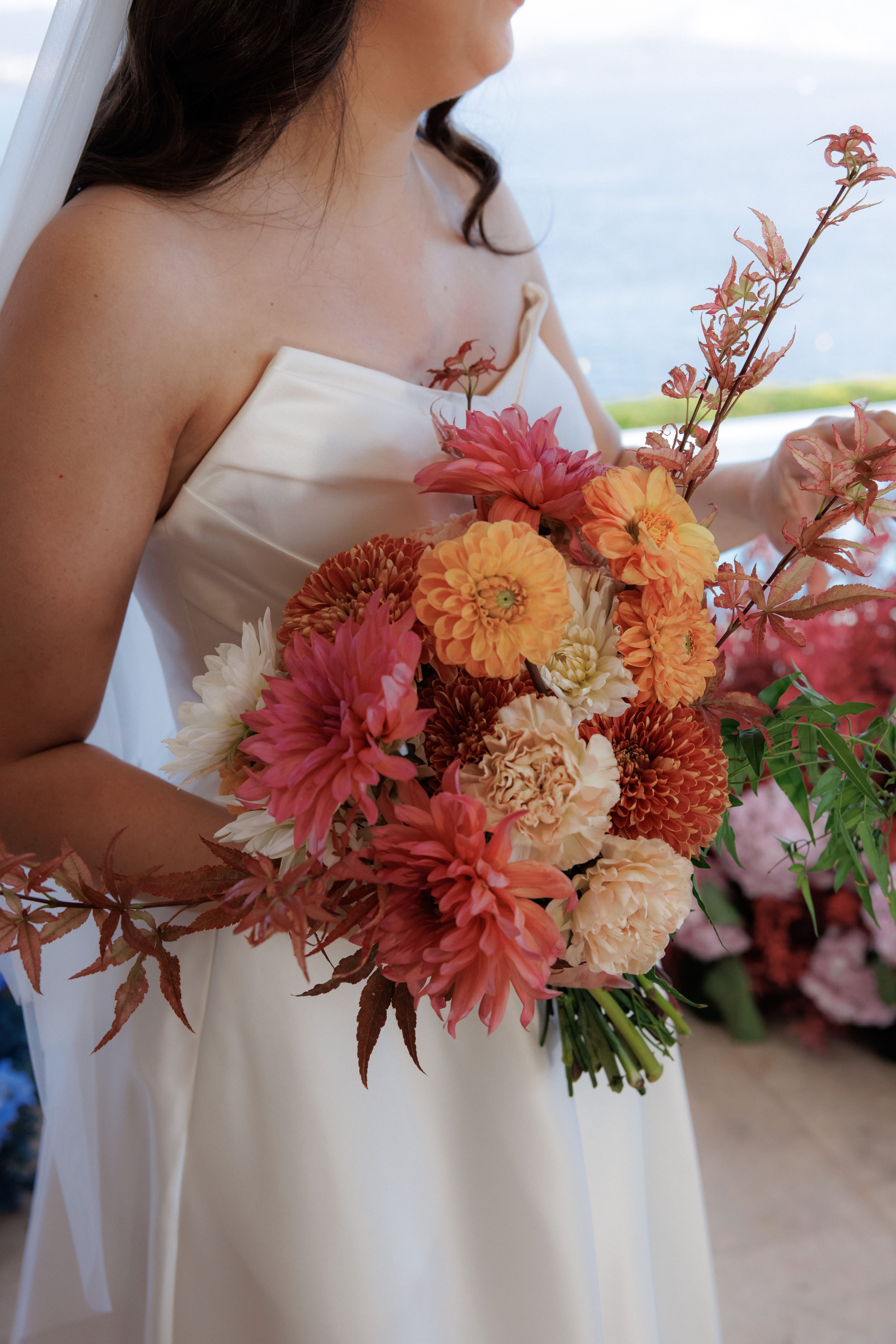 A rounded bridal bouquet of dahlias in coral, rust, apricot and peach, with cream chrysanthemums and peach garden roses, held against a white strapless satin gown. Trailing sprays of red-tipped Japanese maple leaves and green foliage frame the arrangement. The wedding flowers were made for Grace and Tristan at Island Athens Riviera in Greece, with a soft blue sea view behind.