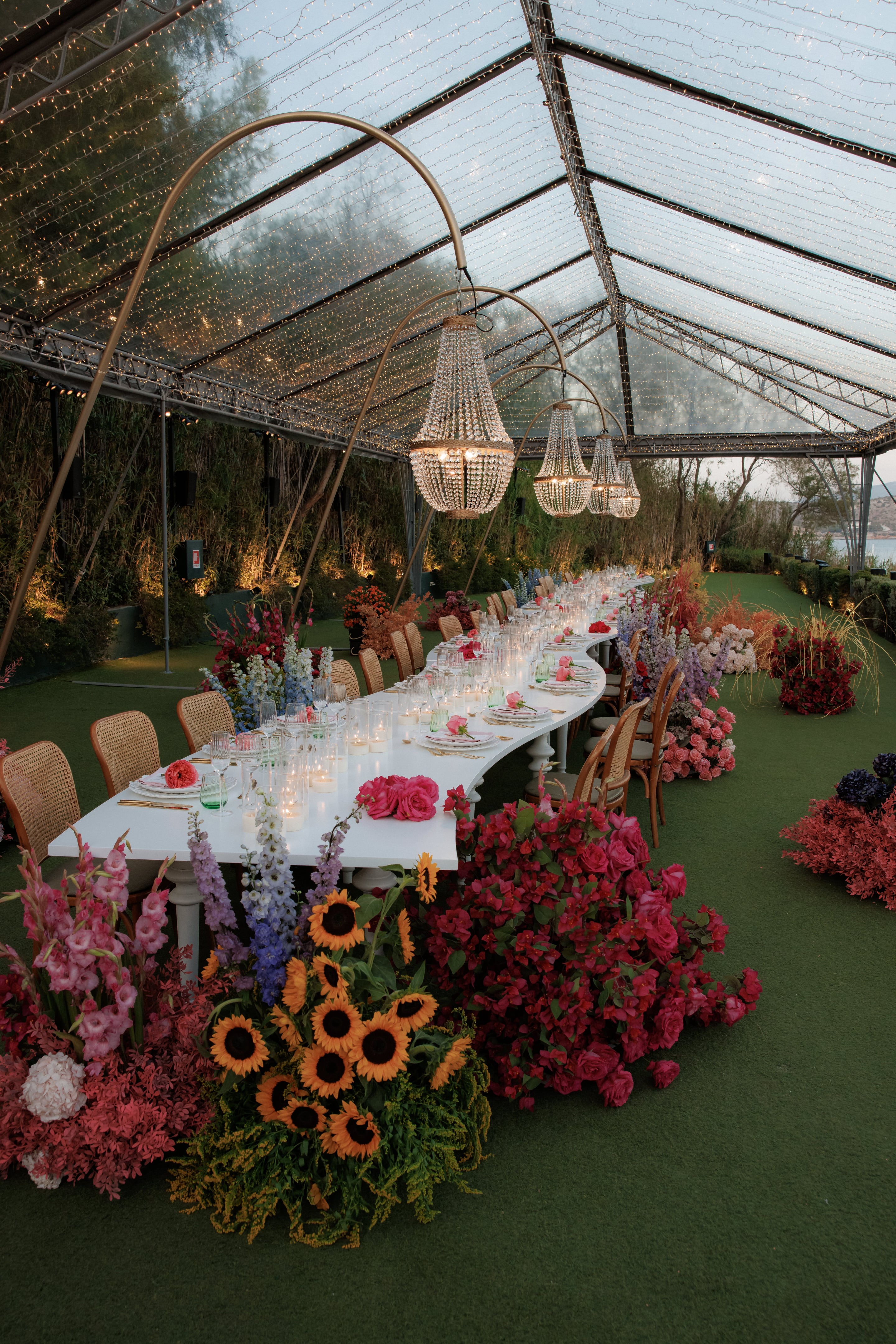 A long, gently curved white banquet table runs beneath a clear, string-lit marquee at Island Athens Riviera in Greece, set for a wedding dinner with candlelit glassware and pink garden roses at each place. Beaded crystal chandeliers hang from arched gold frames overhead, and along the table and beside it stand tall spires of delphinium and lupin in lavender, blue and white, alongside deep red blooms and rose-pink roses. Wicker chairs line the table, with the sea glimpsed through the trees beyond the open walls.