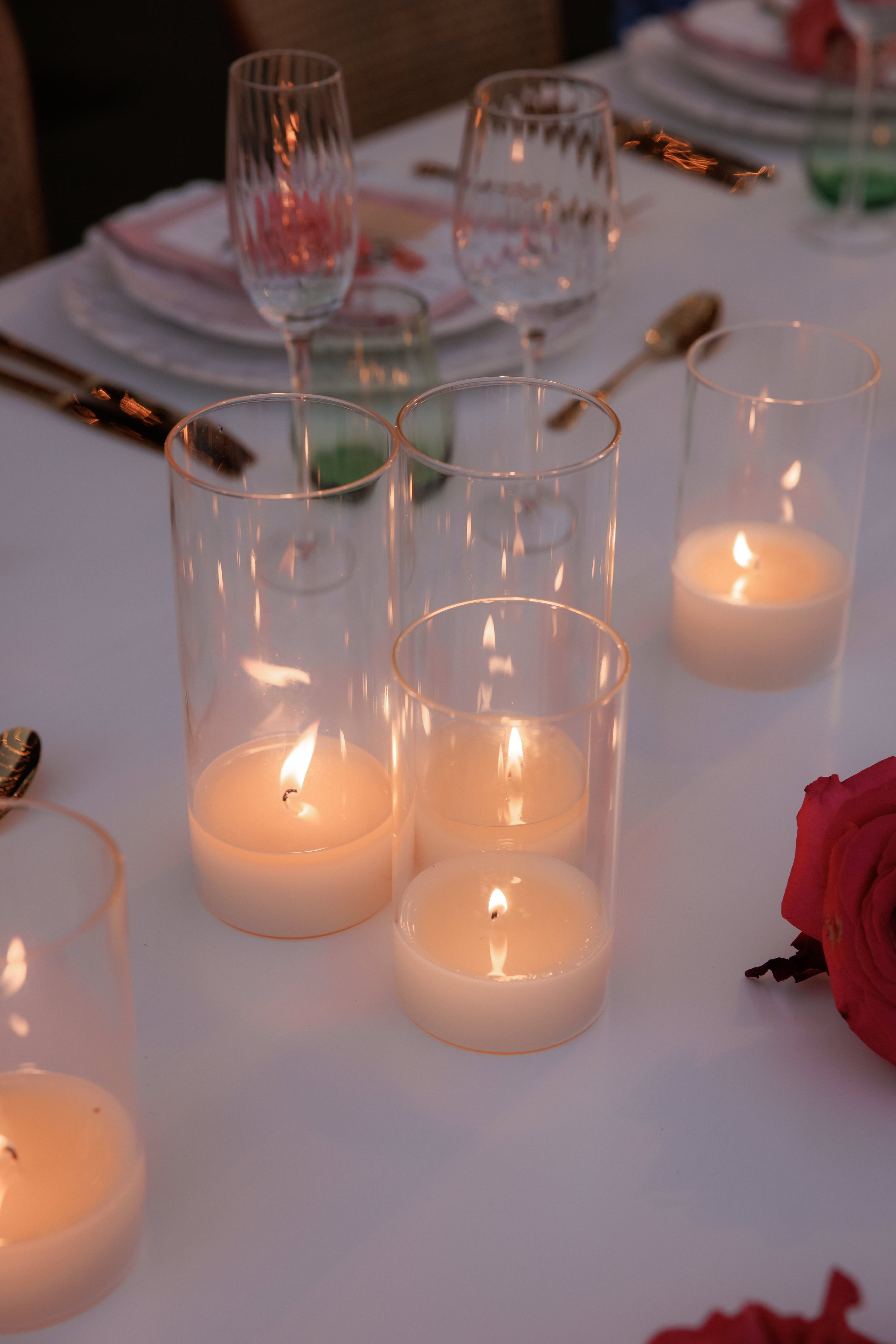 A wedding table setting at Island Athens Riviera in Greece, with three clear glass hurricane candle holders in the foreground, each holding a lit pillar candle whose flames glow warmly. Behind them stand tall fluted champagne flutes and stemless wine glasses on a white linen cloth, with gold cutlery and a pink-bordered plate and napkin. The soft evening light and candle glow give the table an intimate, elegant mood.