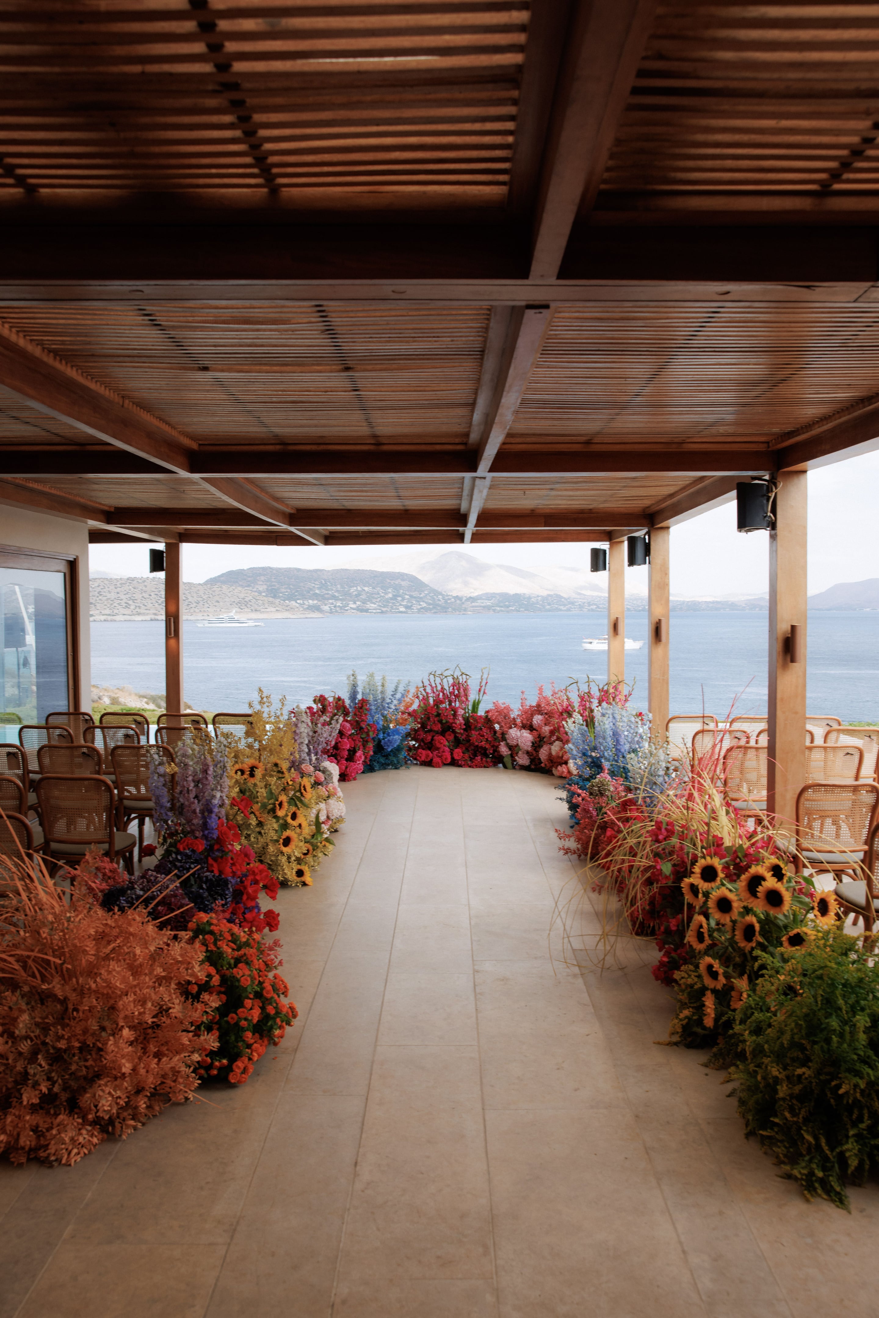 Wedding aisle lined with tall, lush floral borders in red, pink, violet and blue delphinium, with sunflowers and golden grasses along the base, leading between rows of woven cane chairs under a wooden pergola. Beyond the open side, calm blue sea and hazy hills frame the view, with a white yacht drifting in the distance. This is a wedding ceremony setting at Island Athens Riviera in Greece.