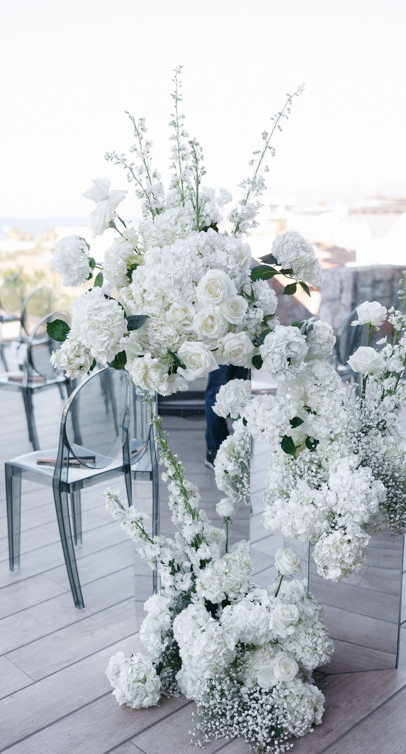 Holly & Mark - In a symphony of elegance and romance, this floral masterpiece frames an intimate moment beneath a brilliant blue sky. The archway, adorned with lush clusters of pristine white blooms and delicate baby's breath, creates a dreamy, ethereal backdrop that captures the essence of sophistication and serenity. The reflective surface beneath adds a touch of modernity, while the gentle ocean breeze seems to whisper promises of a new beginning. This harmonious blend of nature’s finest elements crafts a breathtaking scene, perfect for any discerning couple seeking a timeless and enchanting setting for their special day. - Photo 4