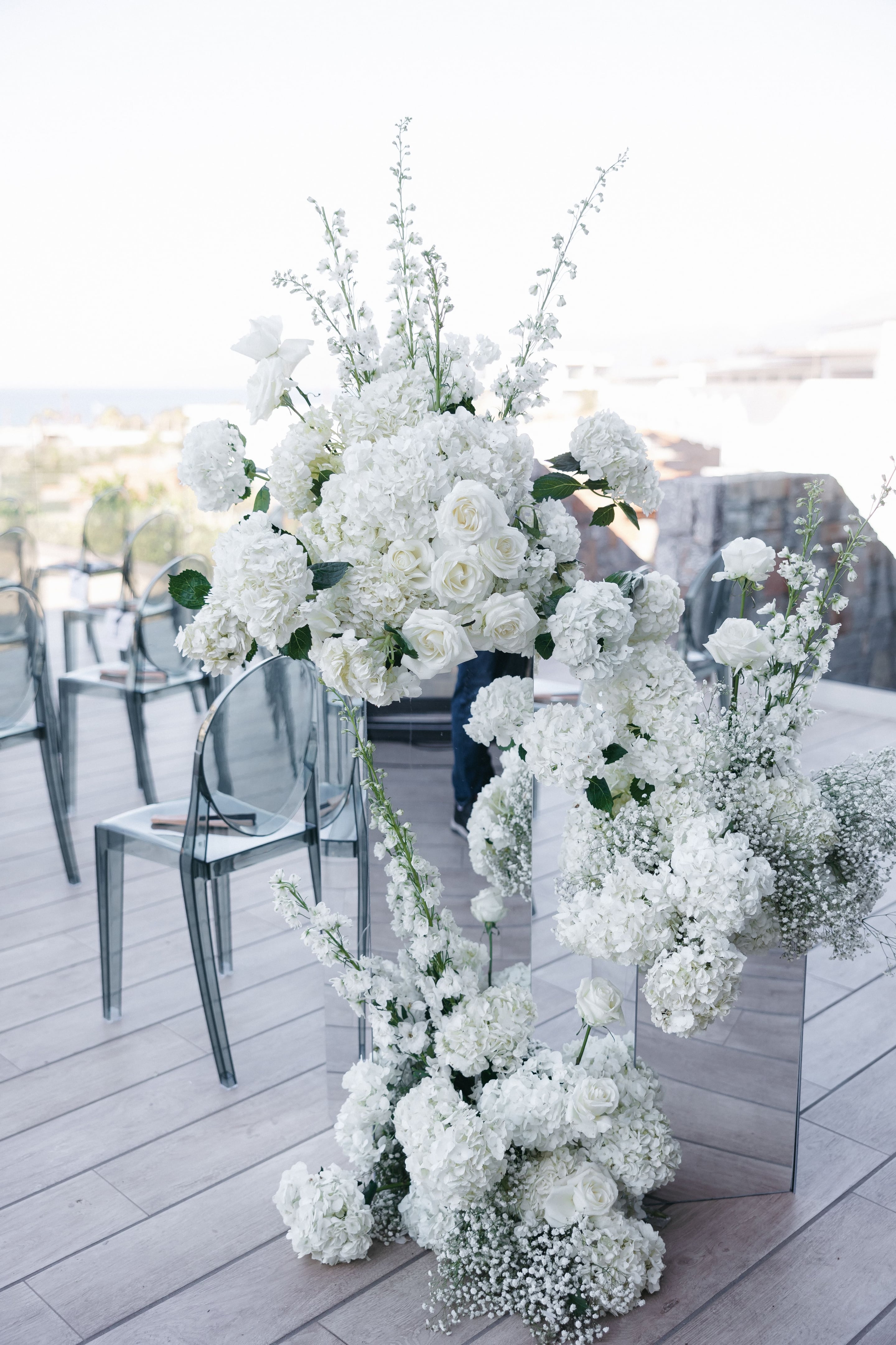 White hydrangeas, white garden roses, stock and wispy gypsophila tumble down two tall mirrored pillars at the wedding ceremony flowers for Holly and Mark at Abaton Hotel in Greece. Behind them, rows of smoky grey transparent chairs line a timber-floored terrace, with a soft blurred sea view under bright daylight. The all-white arrangements, with a few green leaves, give the scene a calm, airy and elegant feel.