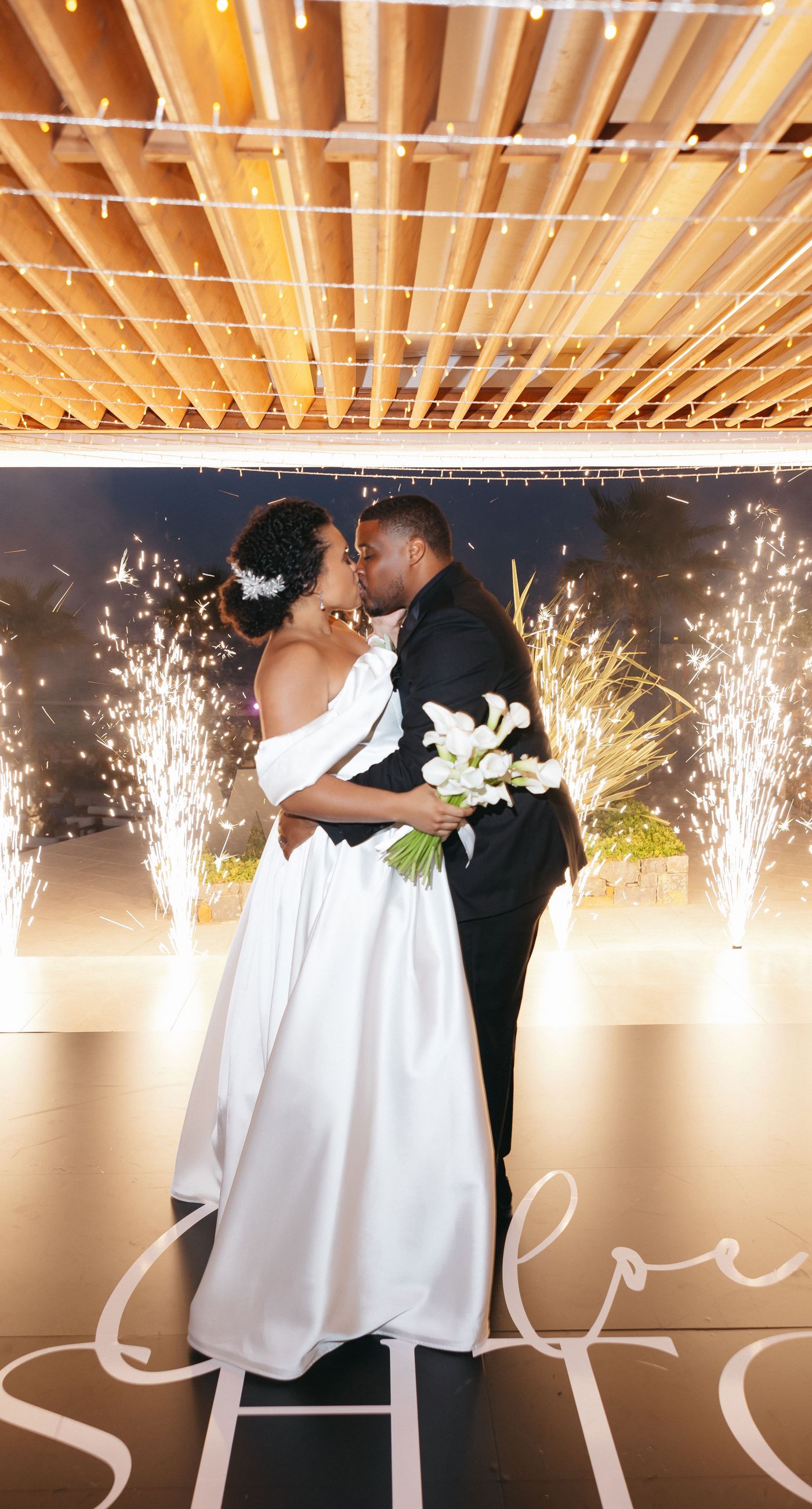 Holly & Mark - In a symphony of elegance and romance, this floral masterpiece frames an intimate moment beneath a brilliant blue sky. The archway, adorned with lush clusters of pristine white blooms and delicate baby's breath, creates a dreamy, ethereal backdrop that captures the essence of sophistication and serenity. The reflective surface beneath adds a touch of modernity, while the gentle ocean breeze seems to whisper promises of a new beginning. This harmonious blend of nature’s finest elements crafts a breathtaking scene, perfect for any discerning couple seeking a timeless and enchanting setting for their special day. - Photo 14