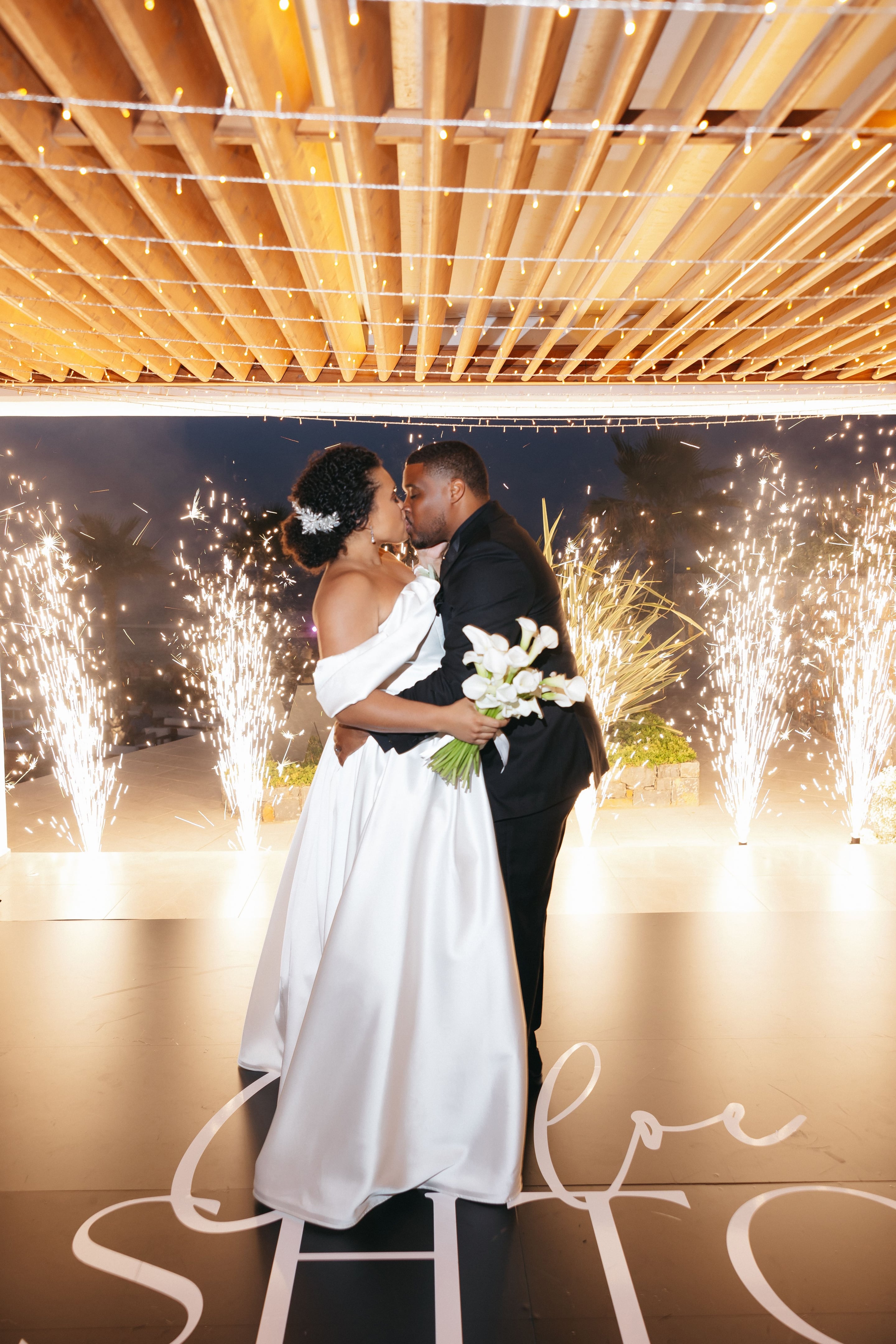 Bride and groom share a kiss at night beneath a wooden pergola strung with fairy lights, while cold fountain sparklers shoot up on either side of them. The bride wears a white satin gown with an off-shoulder sleeve and a pale crystal hair piece, and she holds a bouquet of white calla lilies. The groom is in a black suit, and the golden light and palm silhouettes give the scene a warm, celebratory feel. This is a wedding photograph taken at Abaton Hotel in Greece.
