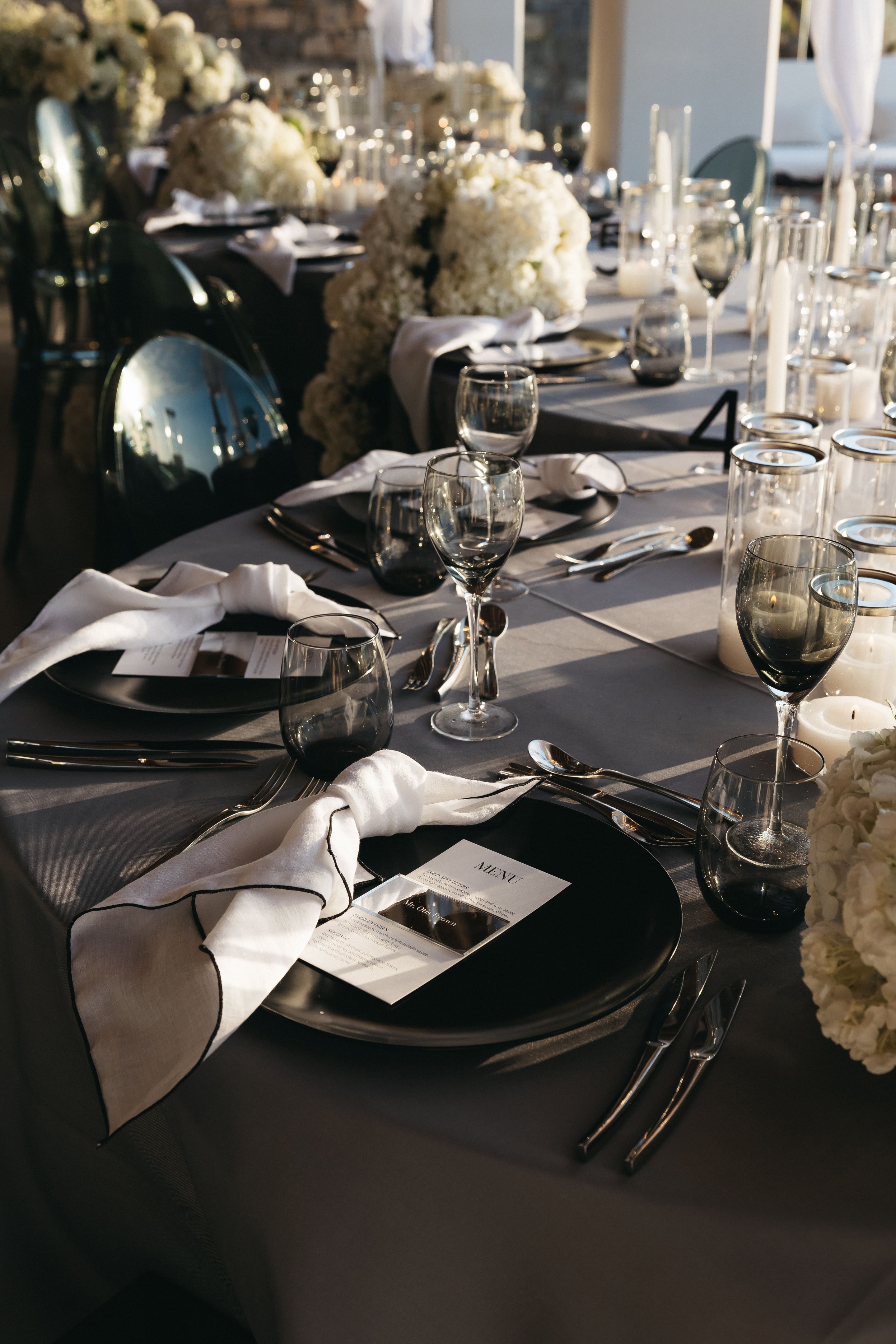 A long wedding table set in monochrome, with grey linen, black charger plates and smoky grey glassware under warm evening light. Clusters of creamy white hydrangeas rise above the place settings, flanked by pillar candles in glass hurricanes with silver rims, and a printed menu card sits on a folded white napkin edged in black stitching. This is a wedding table at the Abaton Hotel in Greece, styled with wedding flowers.