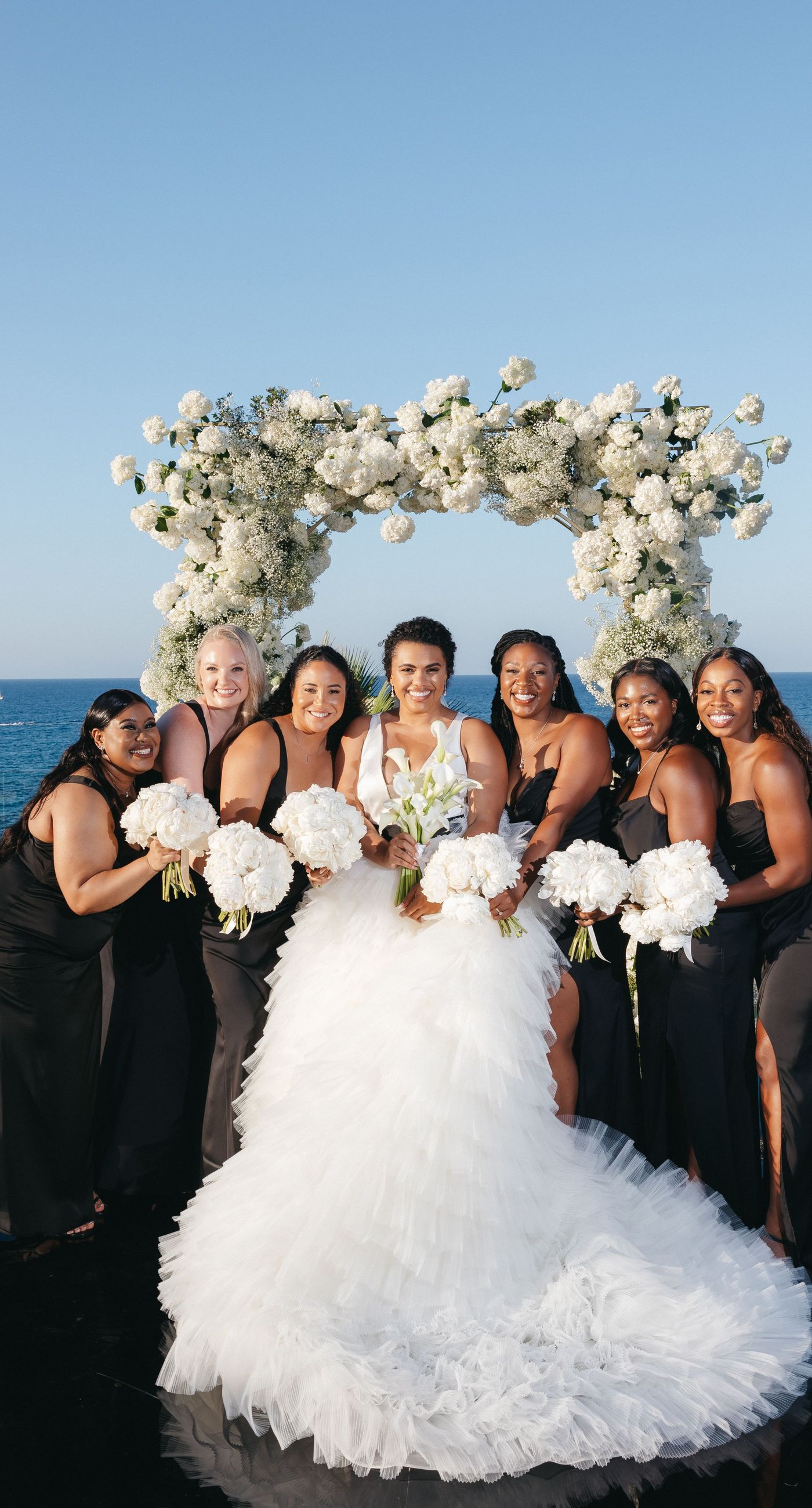 Holly & Mark - In a symphony of elegance and romance, this floral masterpiece frames an intimate moment beneath a brilliant blue sky. The archway, adorned with lush clusters of pristine white blooms and delicate baby's breath, creates a dreamy, ethereal backdrop that captures the essence of sophistication and serenity. The reflective surface beneath adds a touch of modernity, while the gentle ocean breeze seems to whisper promises of a new beginning. This harmonious blend of nature’s finest elements crafts a breathtaking scene, perfect for any discerning couple seeking a timeless and enchanting setting for their special day. - Photo 2