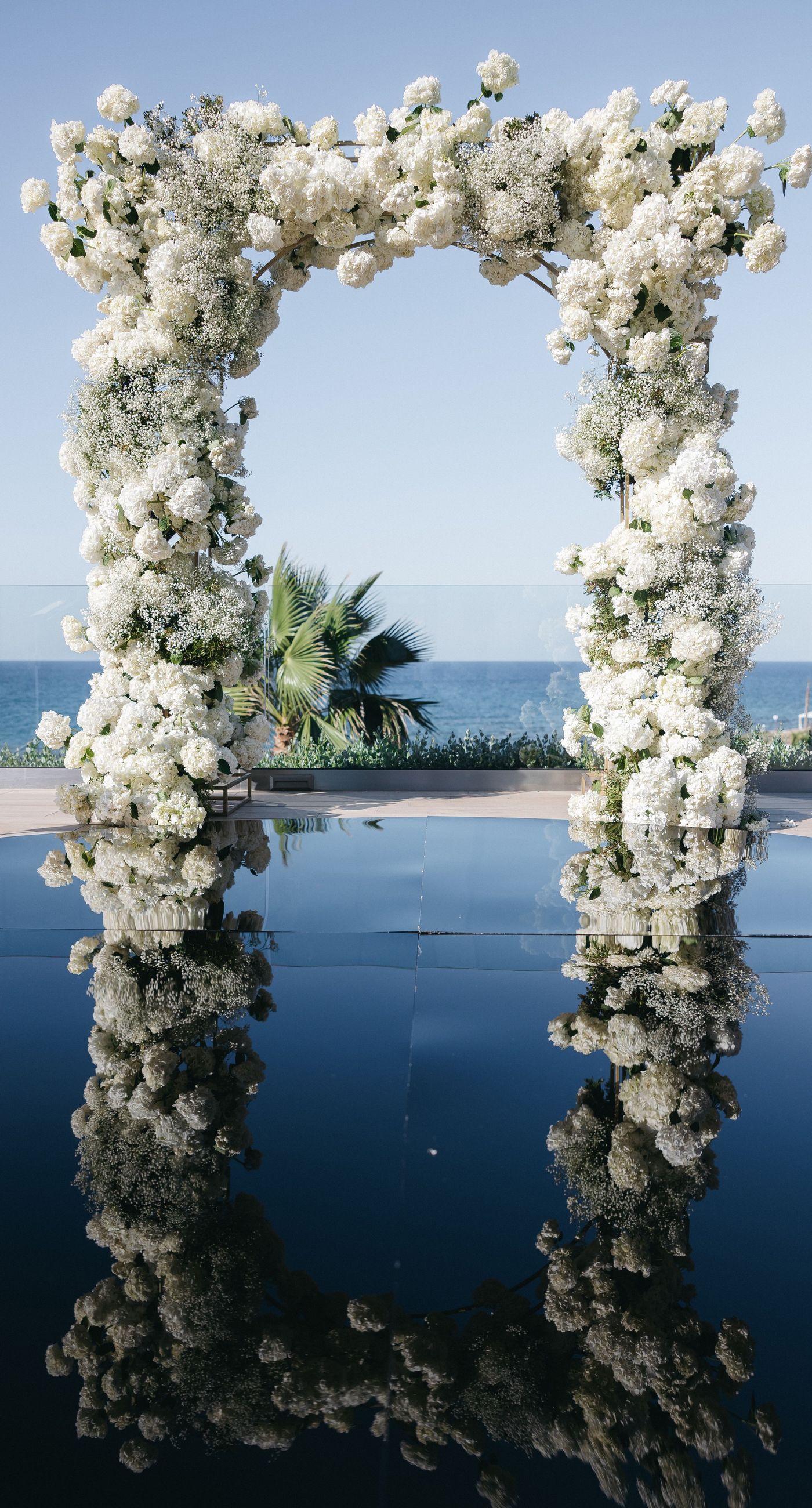 Holly & Mark - In a symphony of elegance and romance, this floral masterpiece frames an intimate moment beneath a brilliant blue sky. The archway, adorned with lush clusters of pristine white blooms and delicate baby's breath, creates a dreamy, ethereal backdrop that captures the essence of sophistication and serenity. The reflective surface beneath adds a touch of modernity, while the gentle ocean breeze seems to whisper promises of a new beginning. This harmonious blend of nature’s finest elements crafts a breathtaking scene, perfect for any discerning couple seeking a timeless and enchanting setting for their special day. - Photo 1