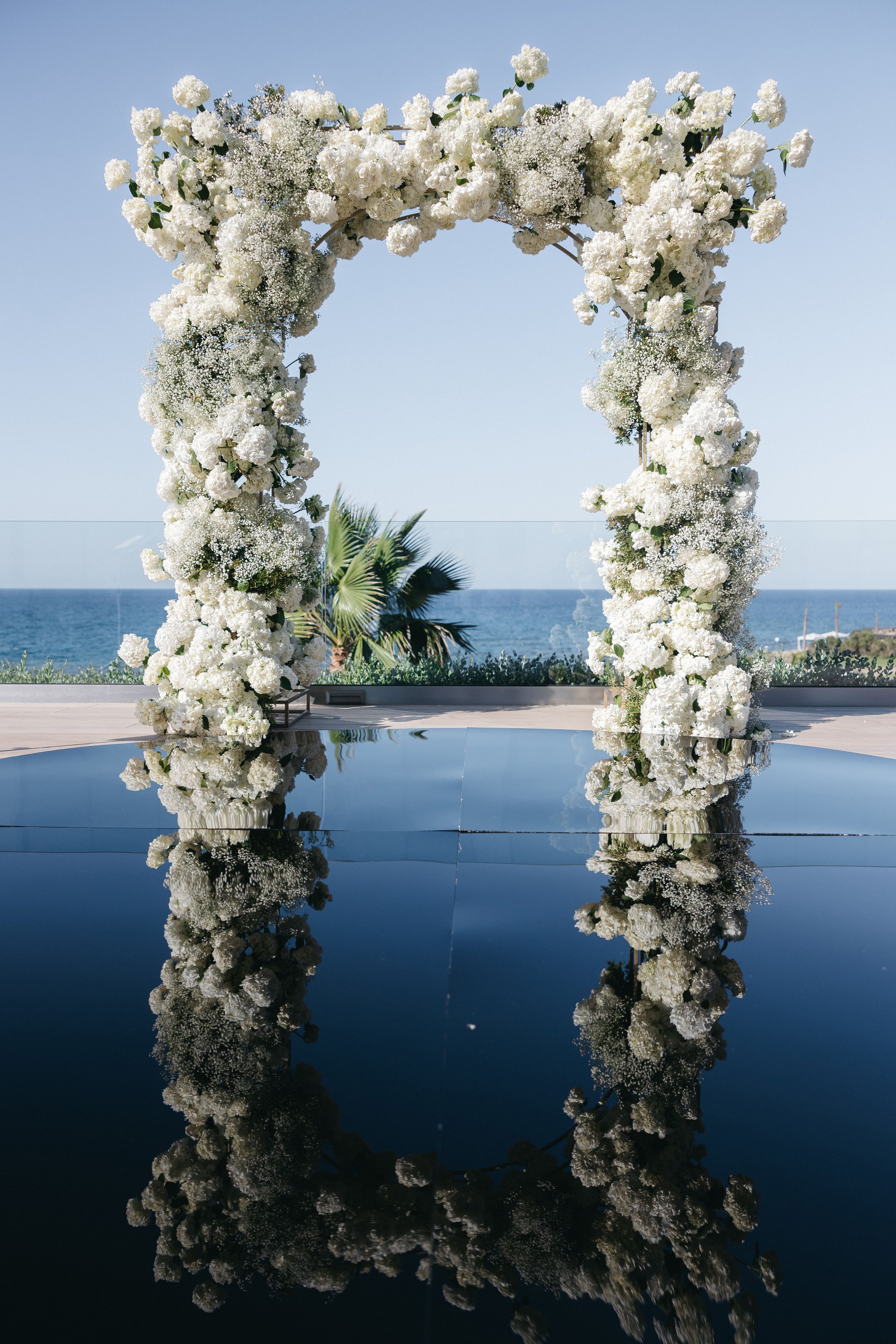 A tall white wedding arch covered in white hydrangeas, roses and airy baby's breath stands beside a still pool of dark blue water, its flowers mirrored perfectly on the surface. Behind it, a glass balustrade frames the calm Aegean Sea under a clear pale sky, with a fan palm at the edge. This wedding flower installation at Abaton Hotel in Greece is all white and serene.