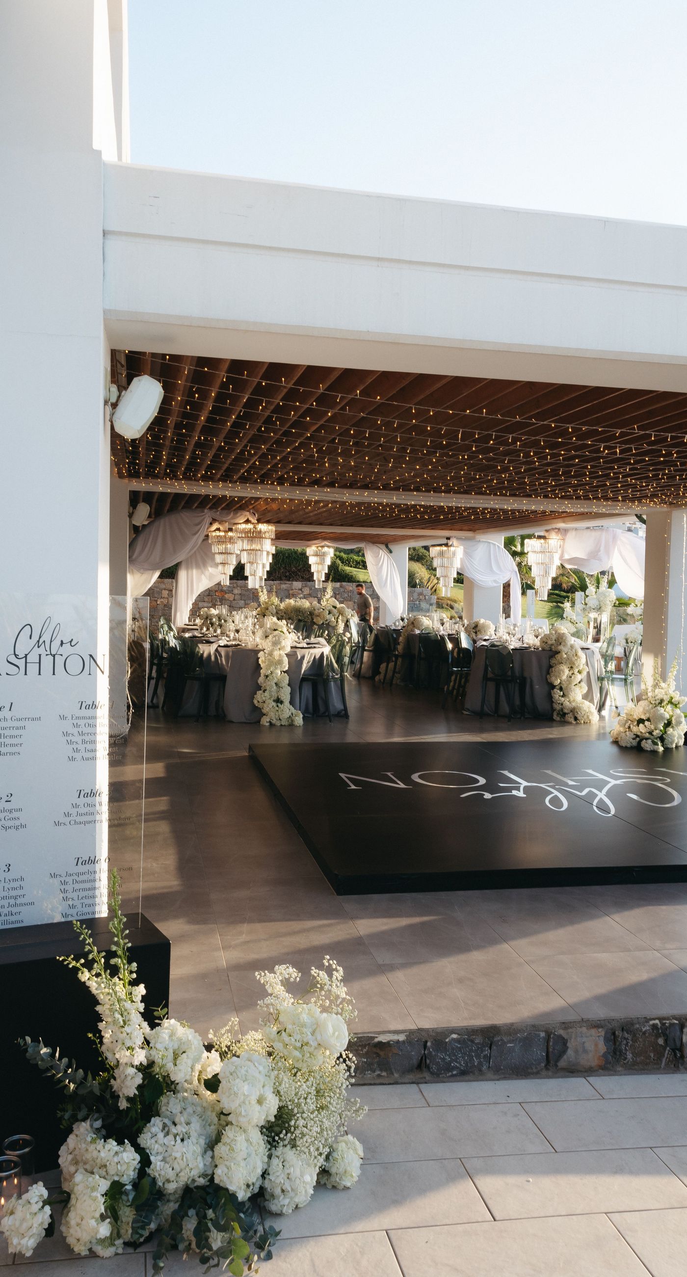 Holly & Mark - In a symphony of elegance and romance, this floral masterpiece frames an intimate moment beneath a brilliant blue sky. The archway, adorned with lush clusters of pristine white blooms and delicate baby's breath, creates a dreamy, ethereal backdrop that captures the essence of sophistication and serenity. The reflective surface beneath adds a touch of modernity, while the gentle ocean breeze seems to whisper promises of a new beginning. This harmonious blend of nature’s finest elements crafts a breathtaking scene, perfect for any discerning couple seeking a timeless and enchanting setting for their special day. - Photo 12