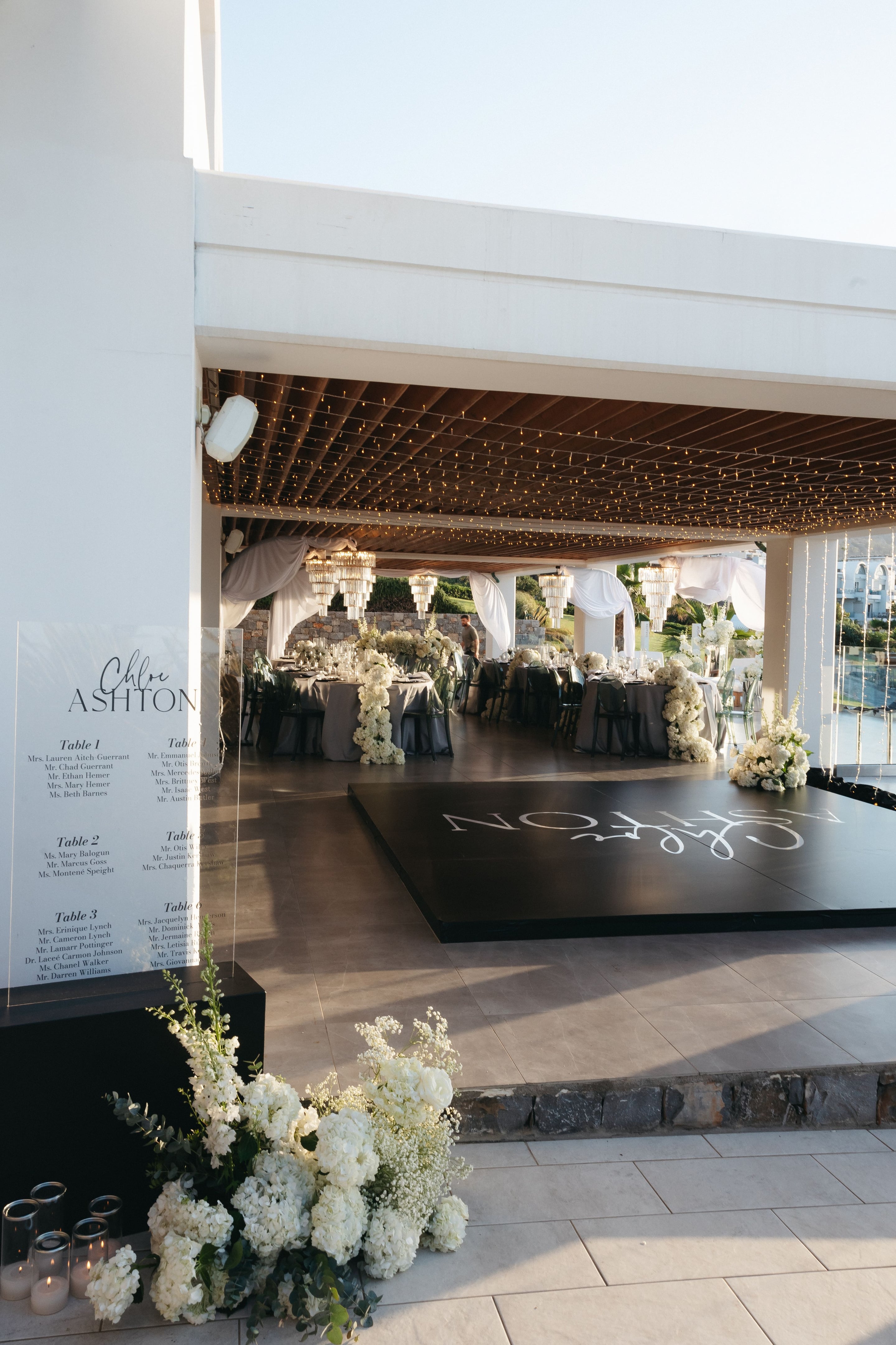 A wedding reception at Abaton Hotel in Greece, under a ceiling strung with warm fairy lights and hung with sheer white drapes. In the foreground, a low arrangement of white hydrangeas, snapdragons, baby's breath and eucalyptus sits beside a clear acrylic seating plan, while a black dance floor bears the couple's names in white script. Round tables dressed in grey linen and set with white floral runners line the room, with crystal chandeliers glowing above.