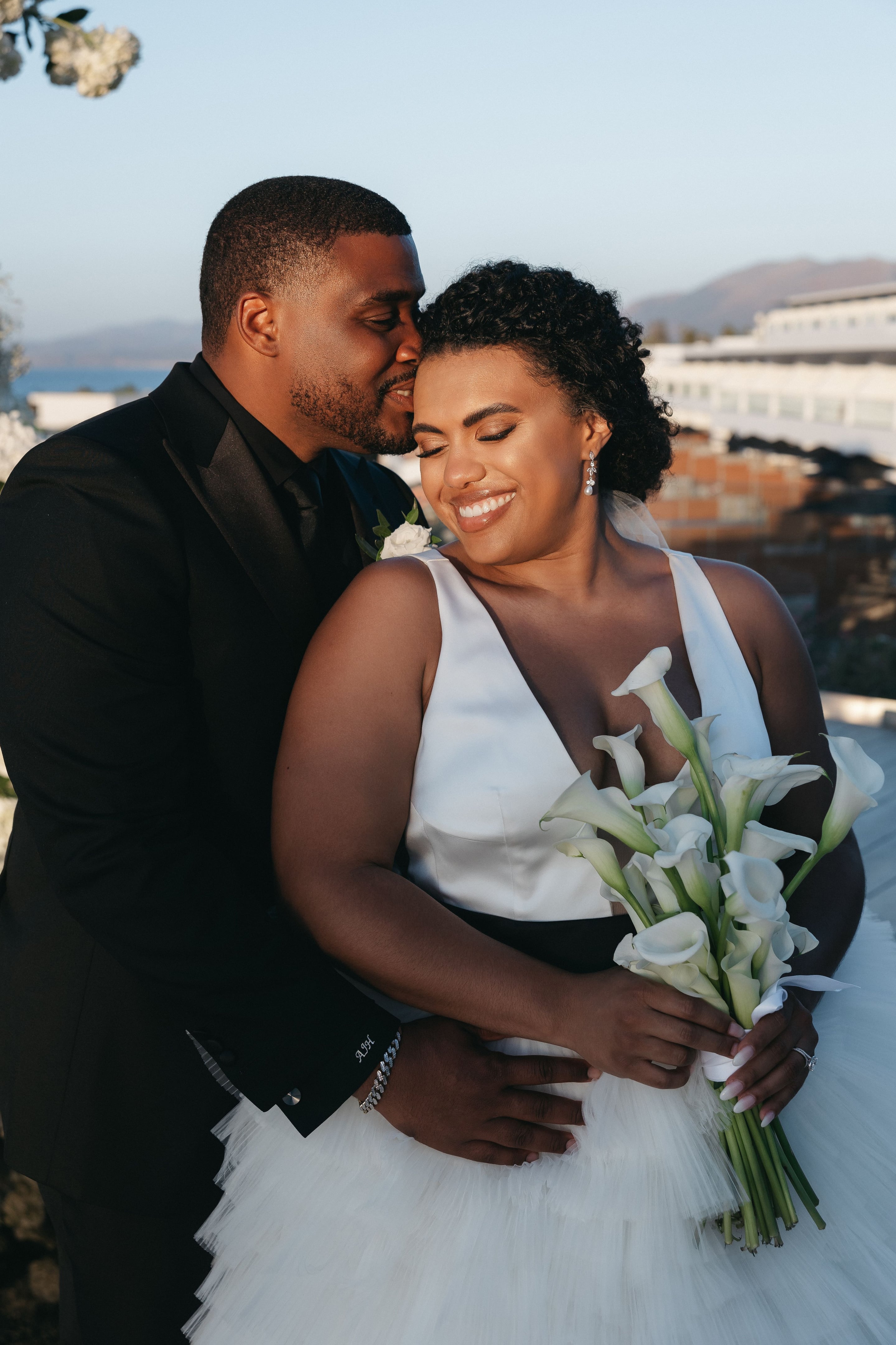 A bride with dark curly hair swept up and a satin white V-neck gown smiles with her eyes closed while a groom in a black suit leans in to kiss her temple. She holds a bouquet of white calla lilies, and a small white rose is pinned to his lapel. This wedding flower photo was taken at Abaton Hotel in Greece, with soft golden evening light, a blurred white building and distant hills behind them.