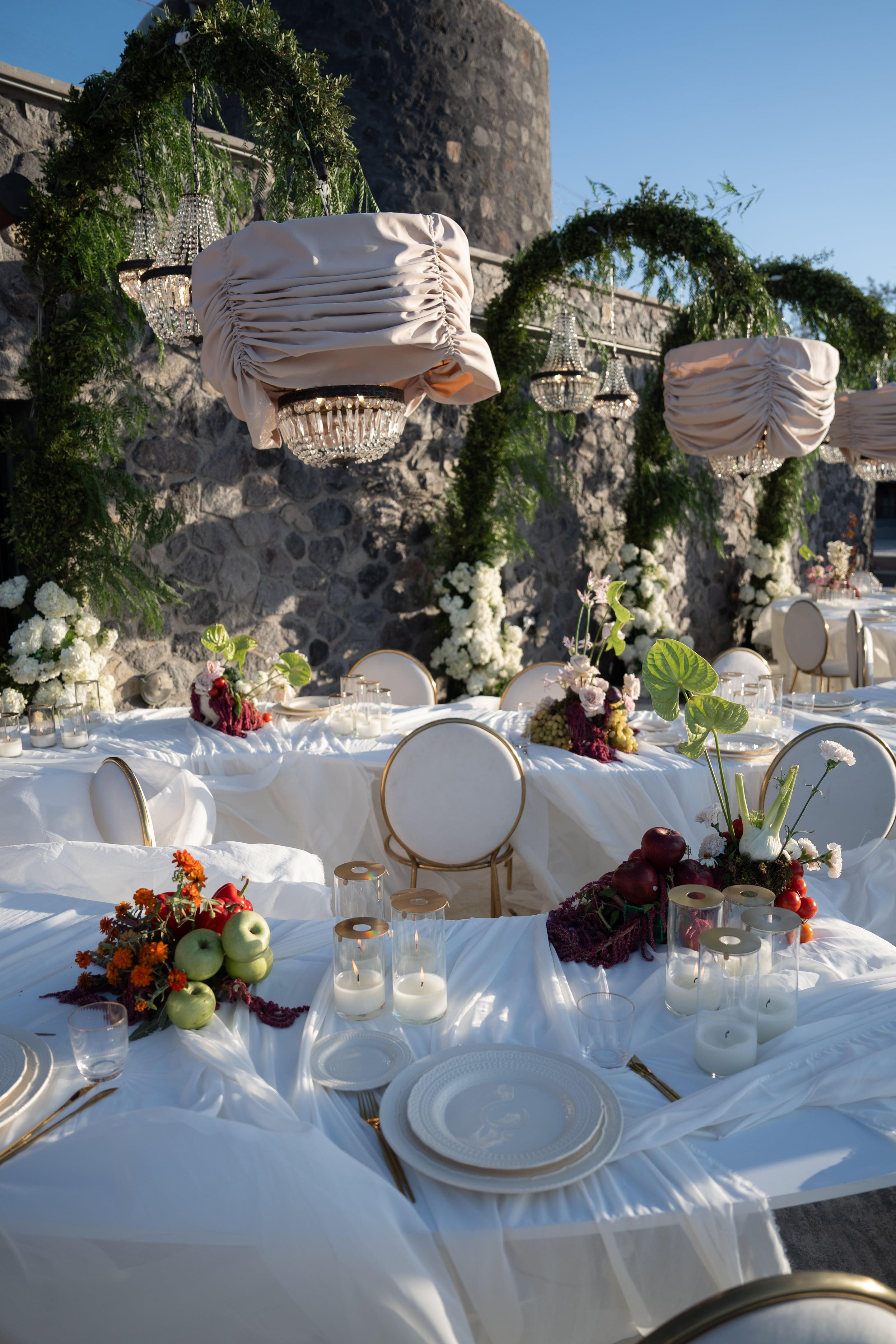 An outdoor wedding reception table dressed in white linen and gold-rimmed chairs, with centrepieces of red apples, green apples, red peppers, tomatoes, orange marigolds and white carnations, plus a few pink blooms. Crystal chandeliers hang beneath draped blush fabric in front of a rough stone wall wreathed with trailing green foliage and white hydrangeas. Warm evening light falls across the scene at Cavo Ventus in Greece, where the wedding flowers and table styling of Isabella and Oliver's celebration create a relaxed yet elegant mood.