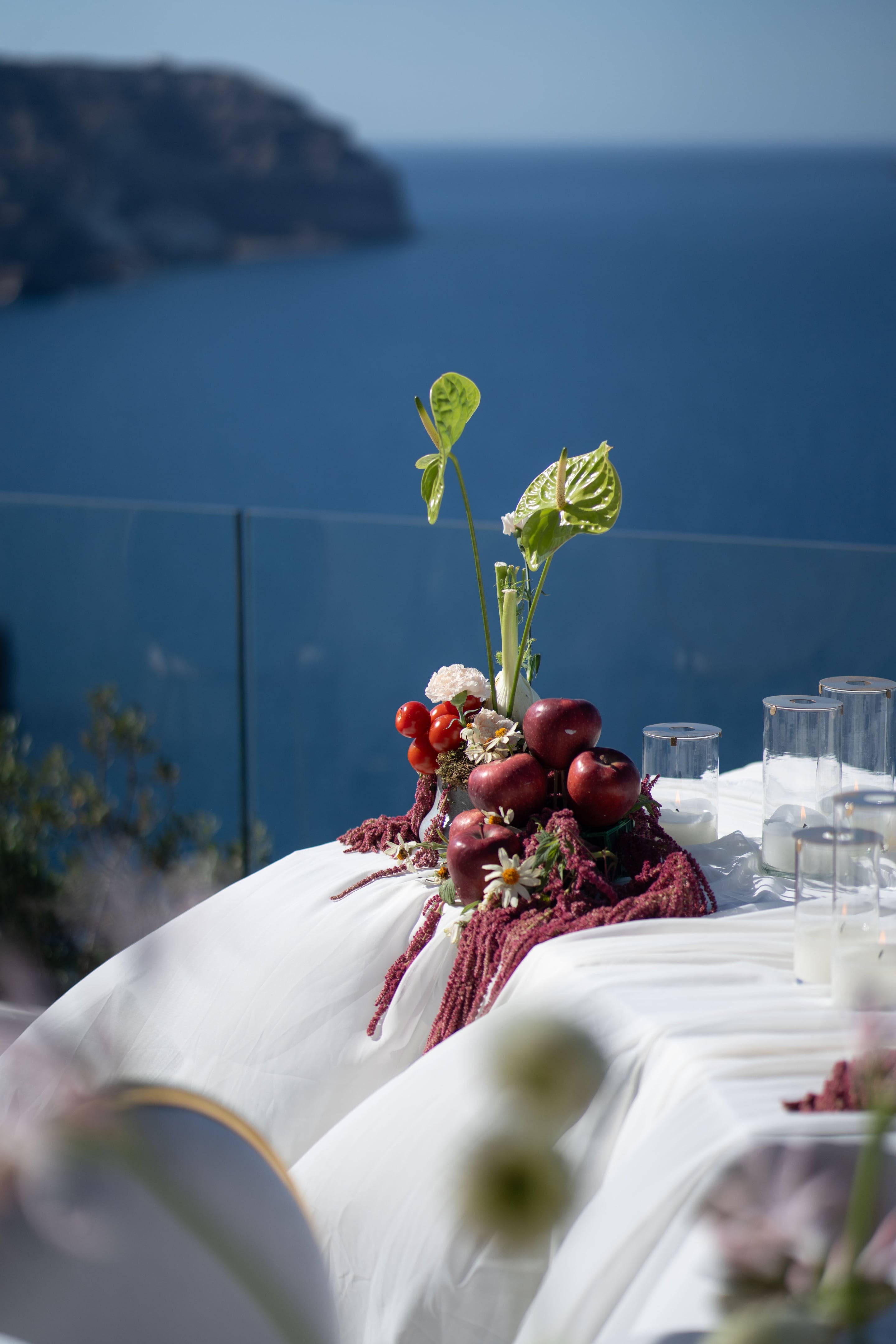A long white-draped wedding table at Cavo Ventus in Greece, centred with a low arrangement of red apples and cherry tomatoes spilling over trailing crimson amaranthus, with white carnations and small daisies tucked between them. Tall glass hurricane candles with lit white pillars line the table to the right, and the blurred blue wall and olive foliage in the background suggest bright daylight.