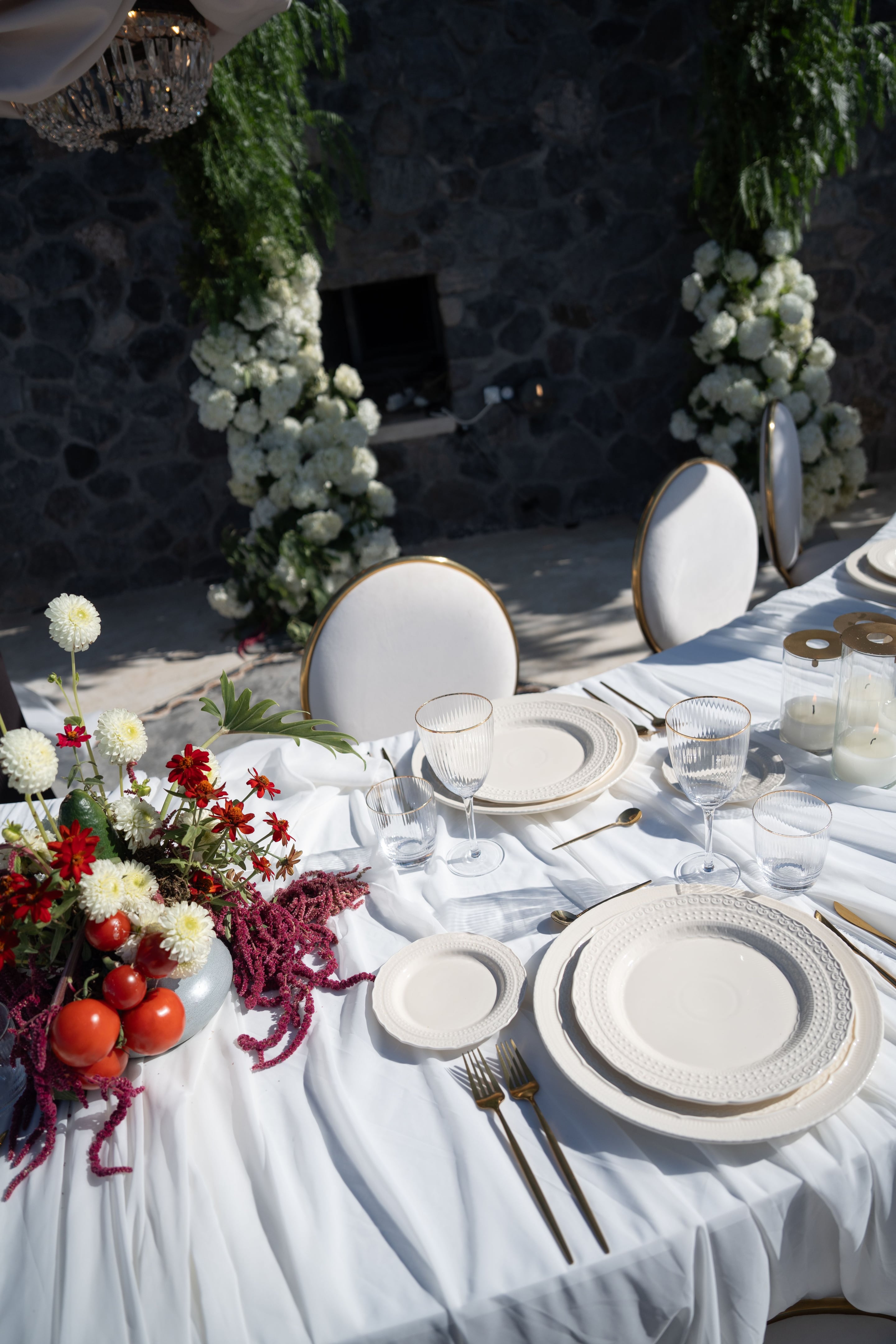 A wedding table set outdoors on flowing white linen, with cream textured plates, gold-rimmed glassware and gold cutlery laid for a place setting. At the left, a low arrangement of white dahlias, red zinnias, a splash of deep magenta amaranthus and ripe red tomatoes sits in a grey ceramic bowl, with a split-leaf philodendron leaf reaching out. Round white chairs with gold trim frame the table in the sunlight, with a white floral arch behind, in Greece.
