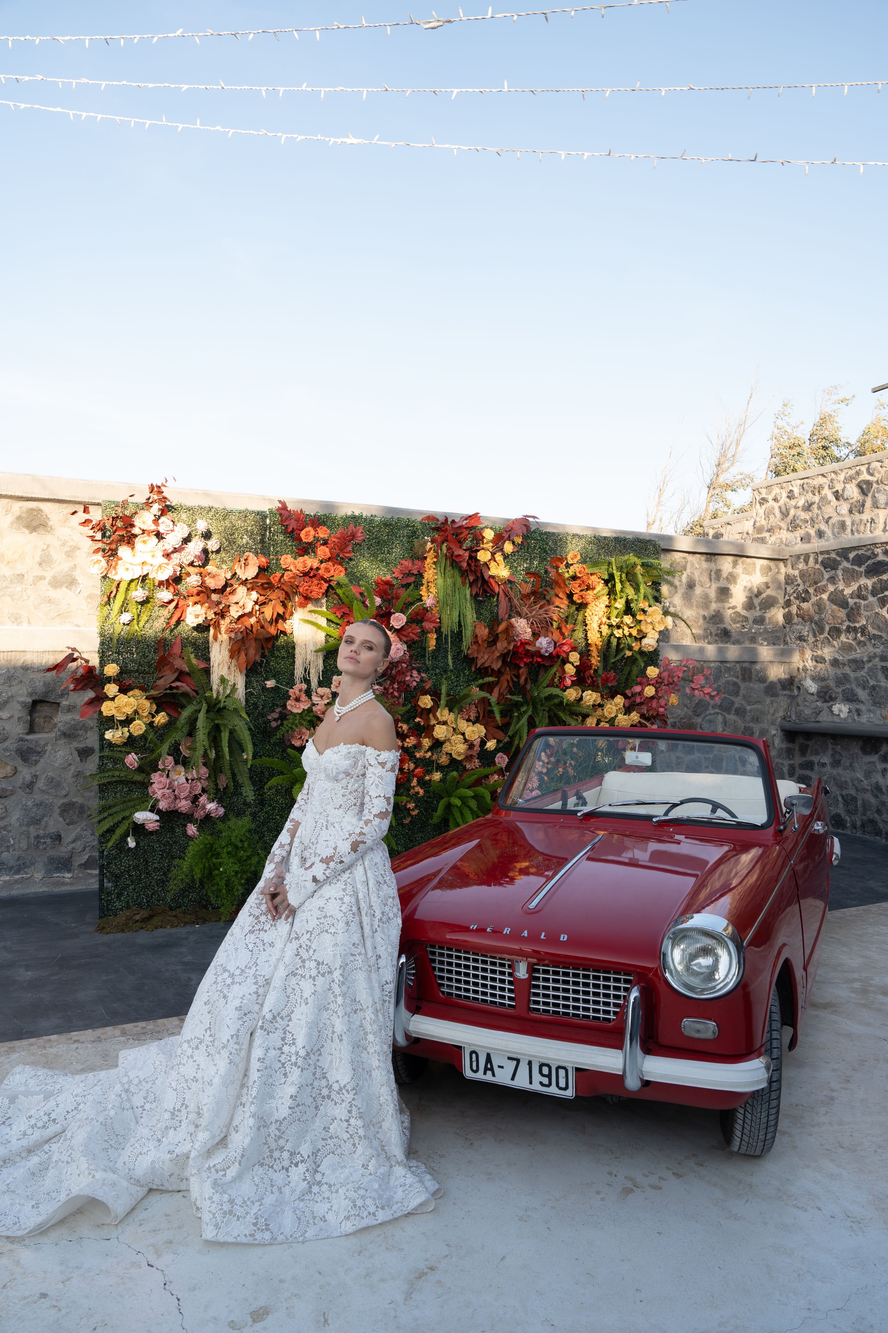 A bride in a long-sleeved, off-the-shoulder lace gown with a sweeping train leans against a vintage red convertible, a Triumph Herald, with a pearl necklace at her throat. Behind her, a tall green wall is covered in autumn-toned blooms: orange roses, cream and peach flowers, golden roses, pink garden roses, and ferns. This wedding flower installation is at Cavo Ventus in Greece, set against weathered stone walls.