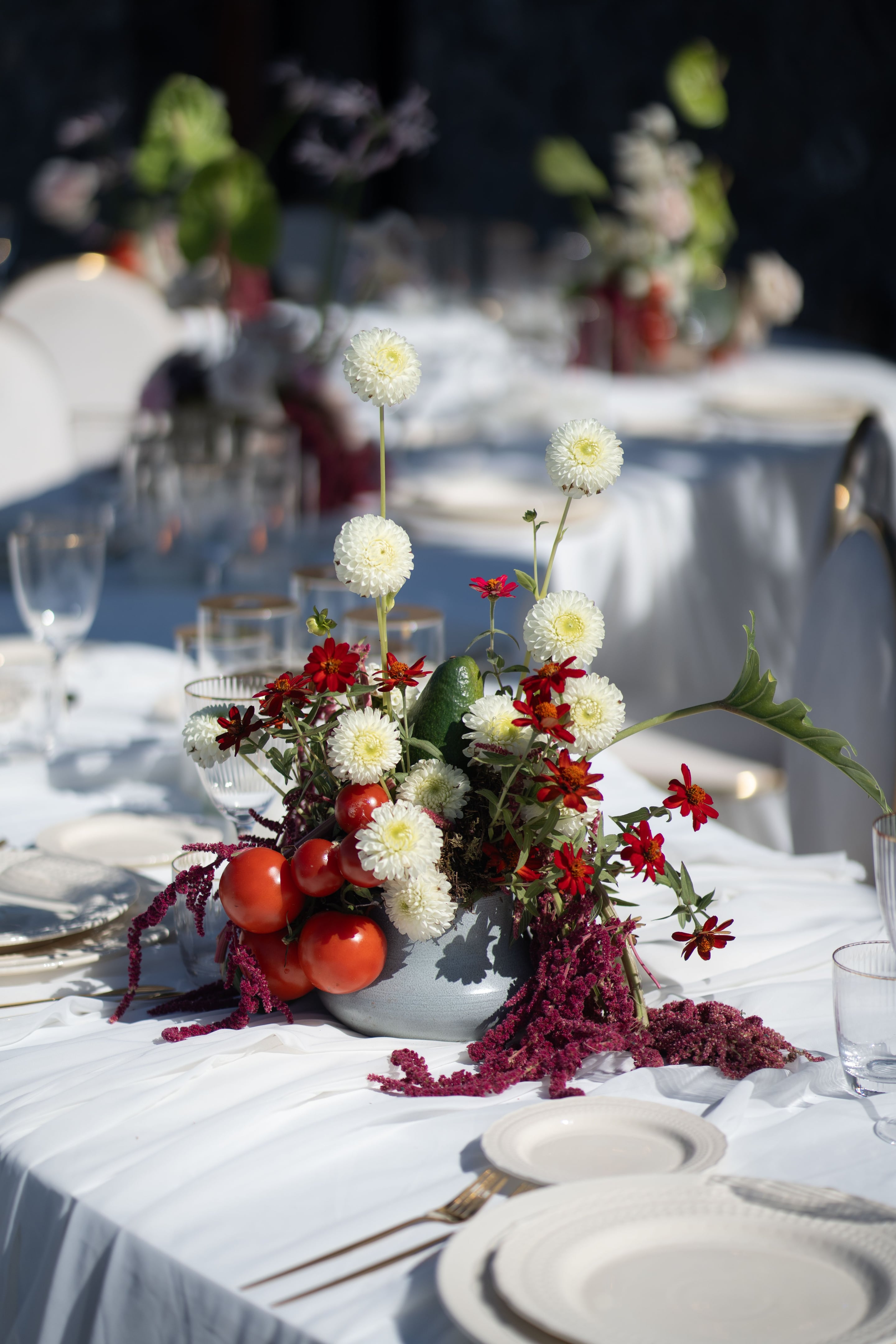 A low grey ceramic bowl centrepiece on a white linen table at Cavo Ventus, Greece, filled with white pompom chrysanthemums, red zinnias and trailing burgundy amaranthus. Tall white blooms rise on bare stems above the arrangement, while cherry tomatoes and a small green gourd sit among the flowers, with a green philodendron-style leaf sweeping out to the right. The wedding table setting, with clear glasses and white plates, glows softly in the sunlight behind it.